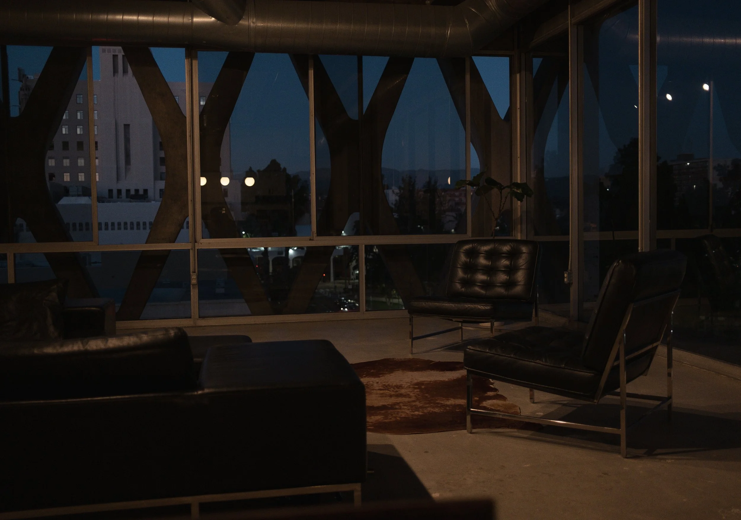 Dimly lit modern interior with black leather chairs, a small table, a potted plant, and large windows showing a cityscape at night with buildings and streetlights.
