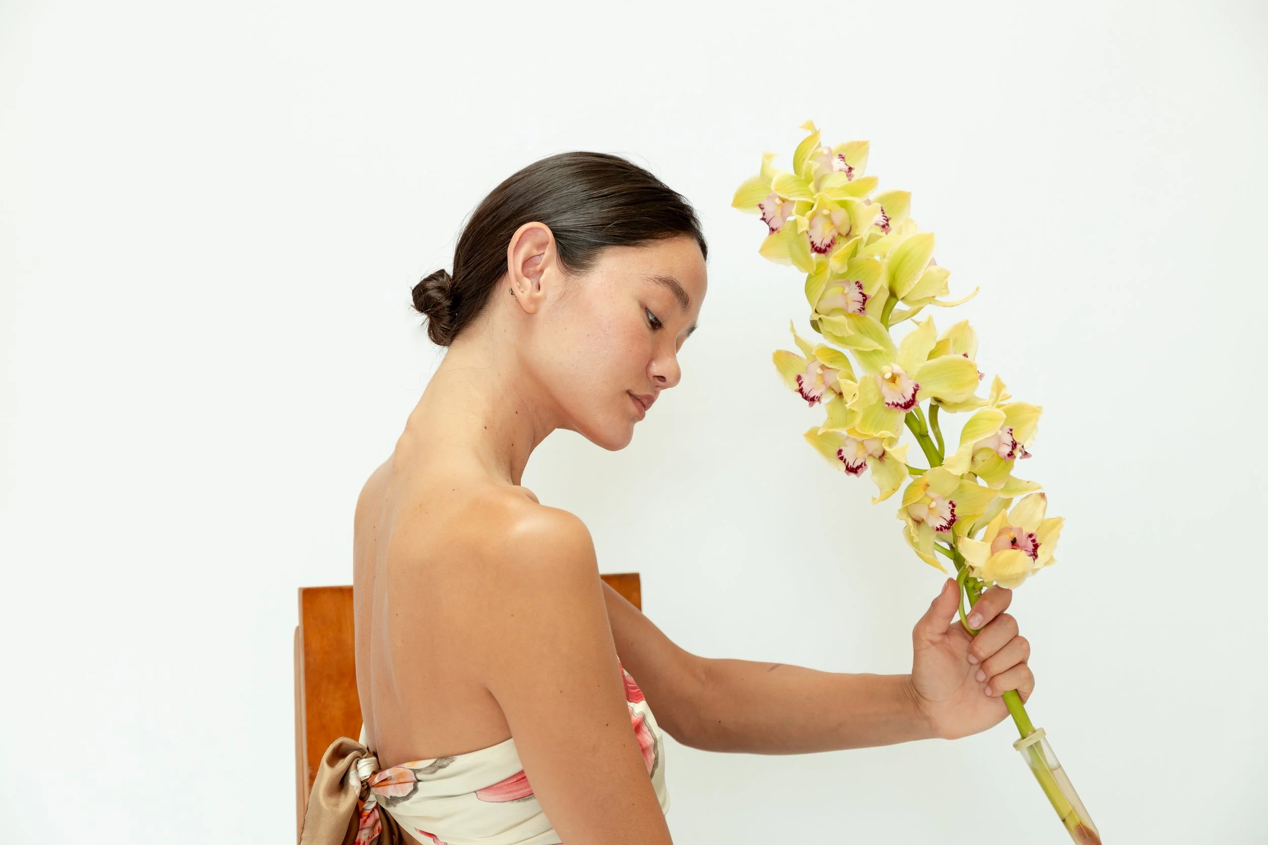 A woman with dark hair tied in a bun, sitting on a wooden chair, holding a large bouquet of yellow orchids with purple centers, wearing a floral dress with the back exposed, against a plain white background.