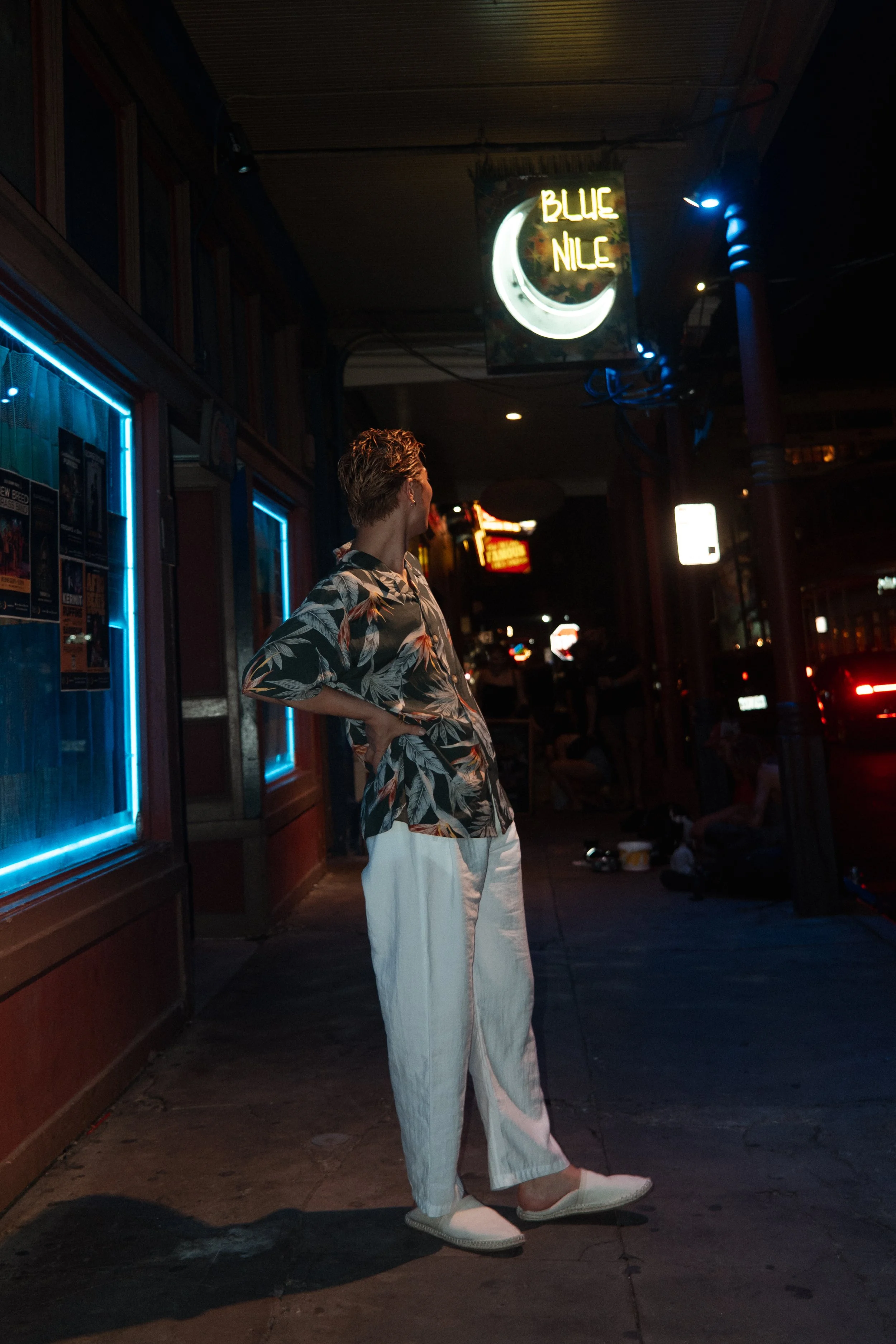A person standing on a city sidewalk at night beneath neon signs, including a sign for Blue Nile, with others in the background, wearing a floral shirt, white pants, and shoes.