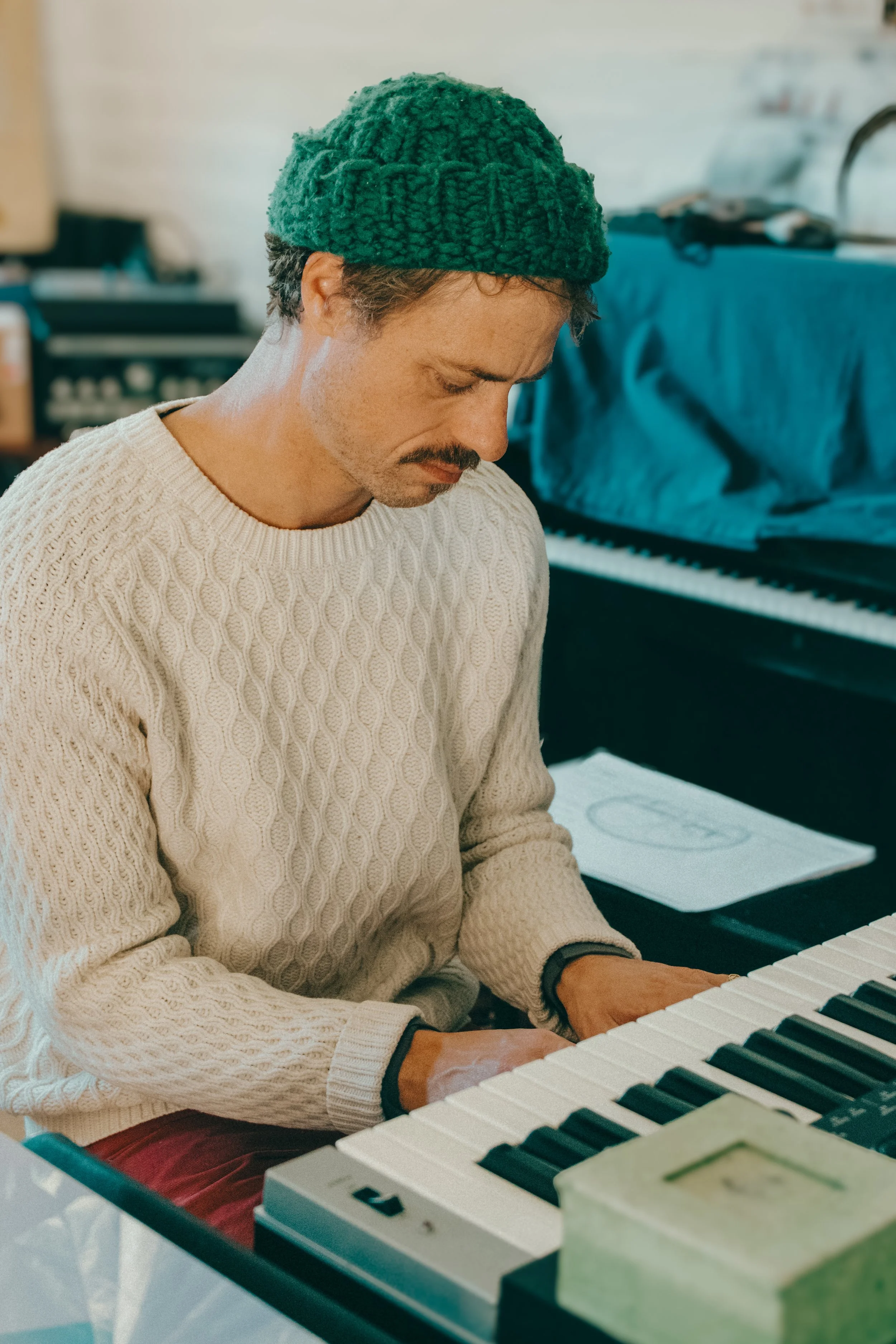 A man wearing a green knit beanie and a cream-colored cable-knit sweater playing a keyboard.