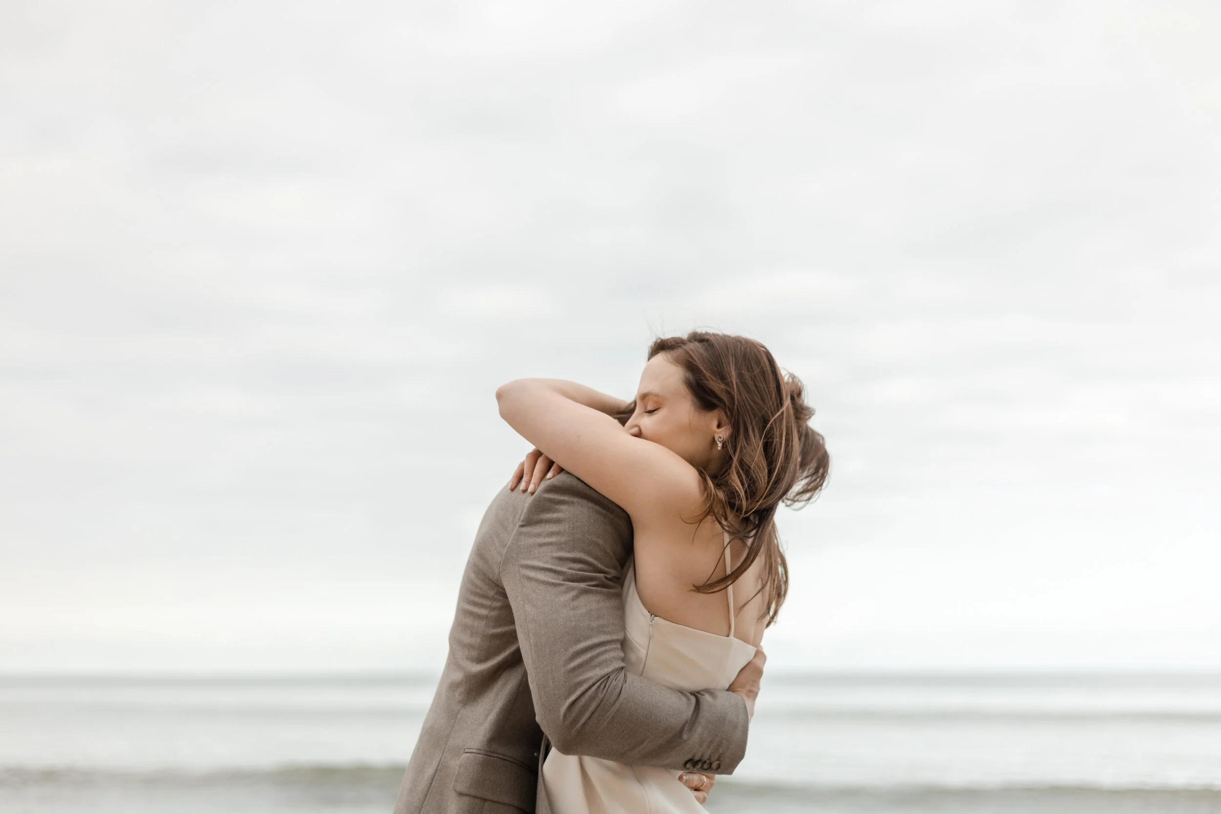 A woman and a man hugging on a beach with cloudy sky.