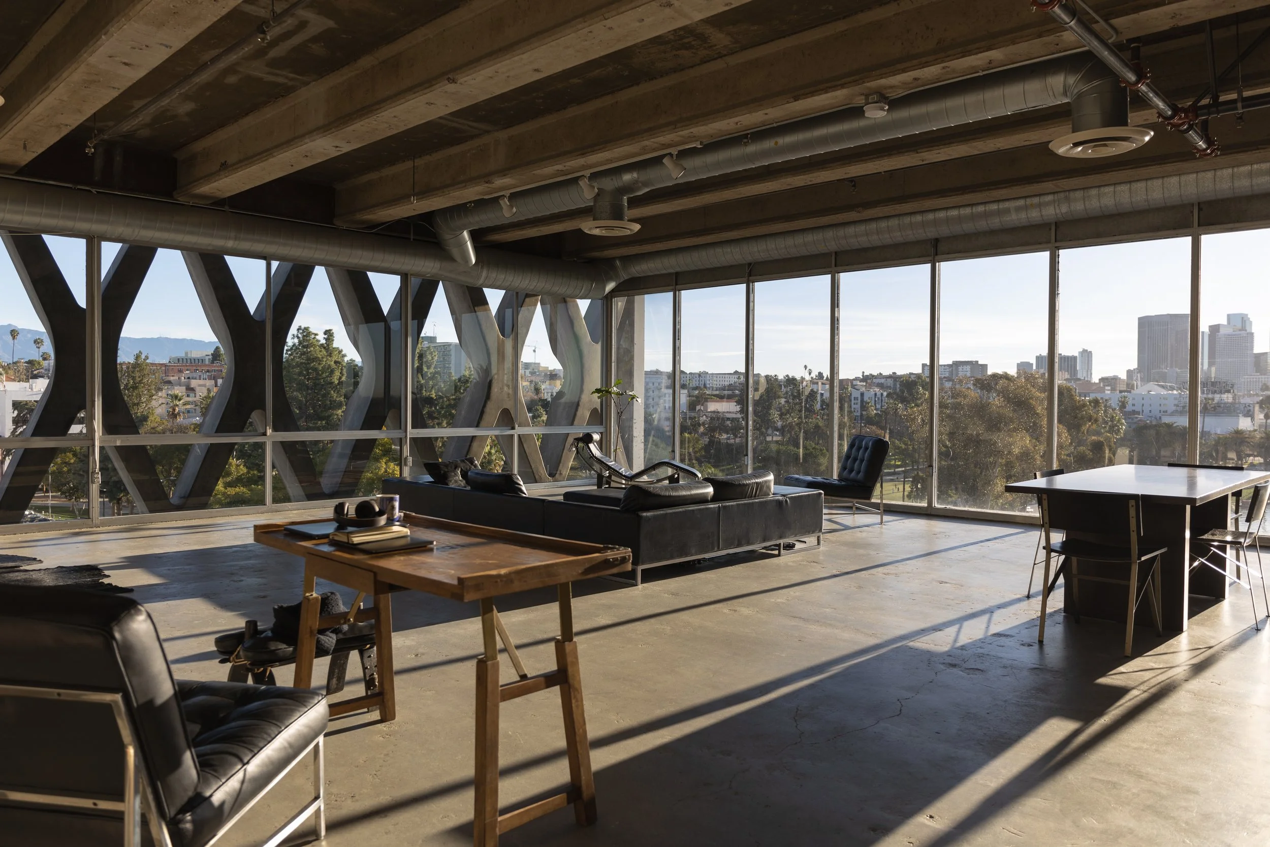 Modern office with large windows overlooking a cityscape, furnished with a black sofa, a chair, a wooden table with books and headphones, and a black table with chairs.