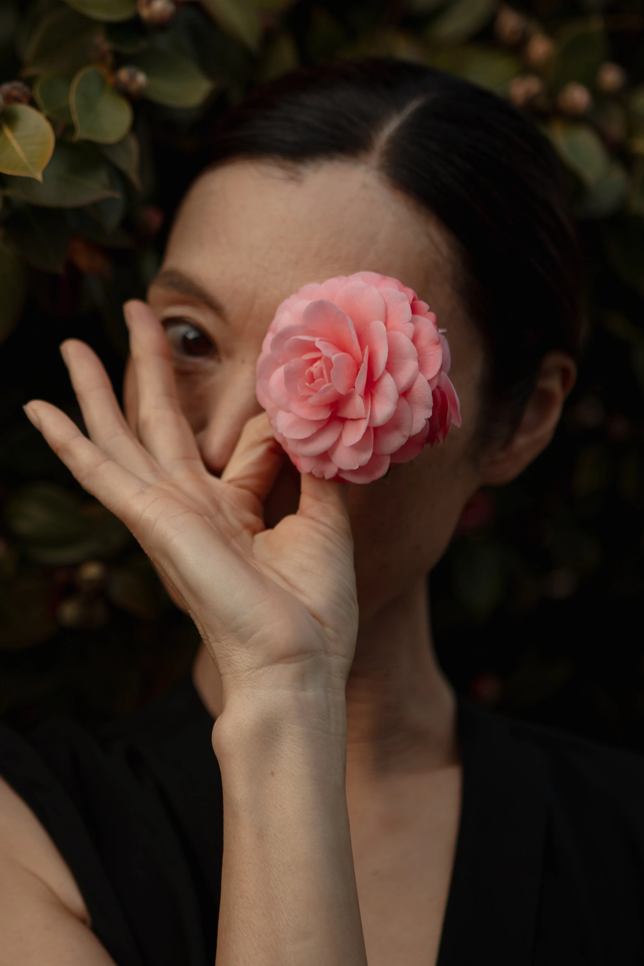 A woman with dark hair holding a pink camellia flower in front of her face, partially covering her eye, with a background of green leaves.