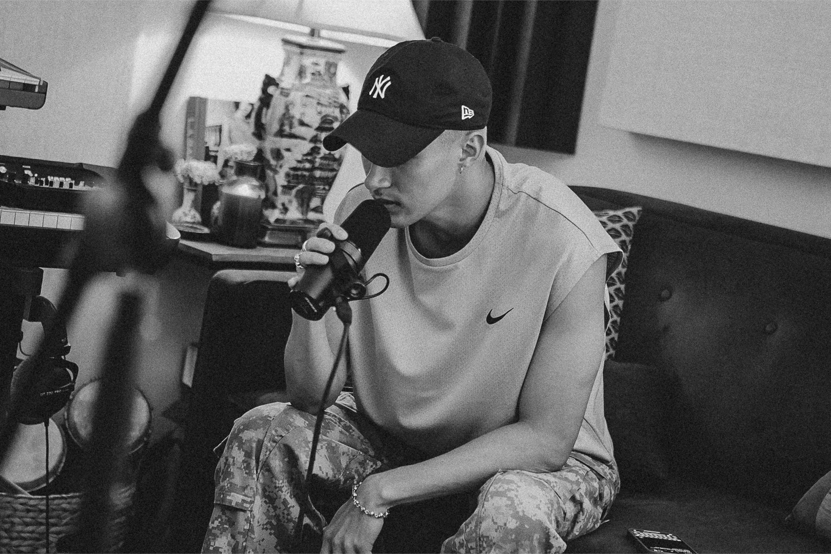 A young man singing into a microphone while sitting on a couch in a room with musical equipment and books.
