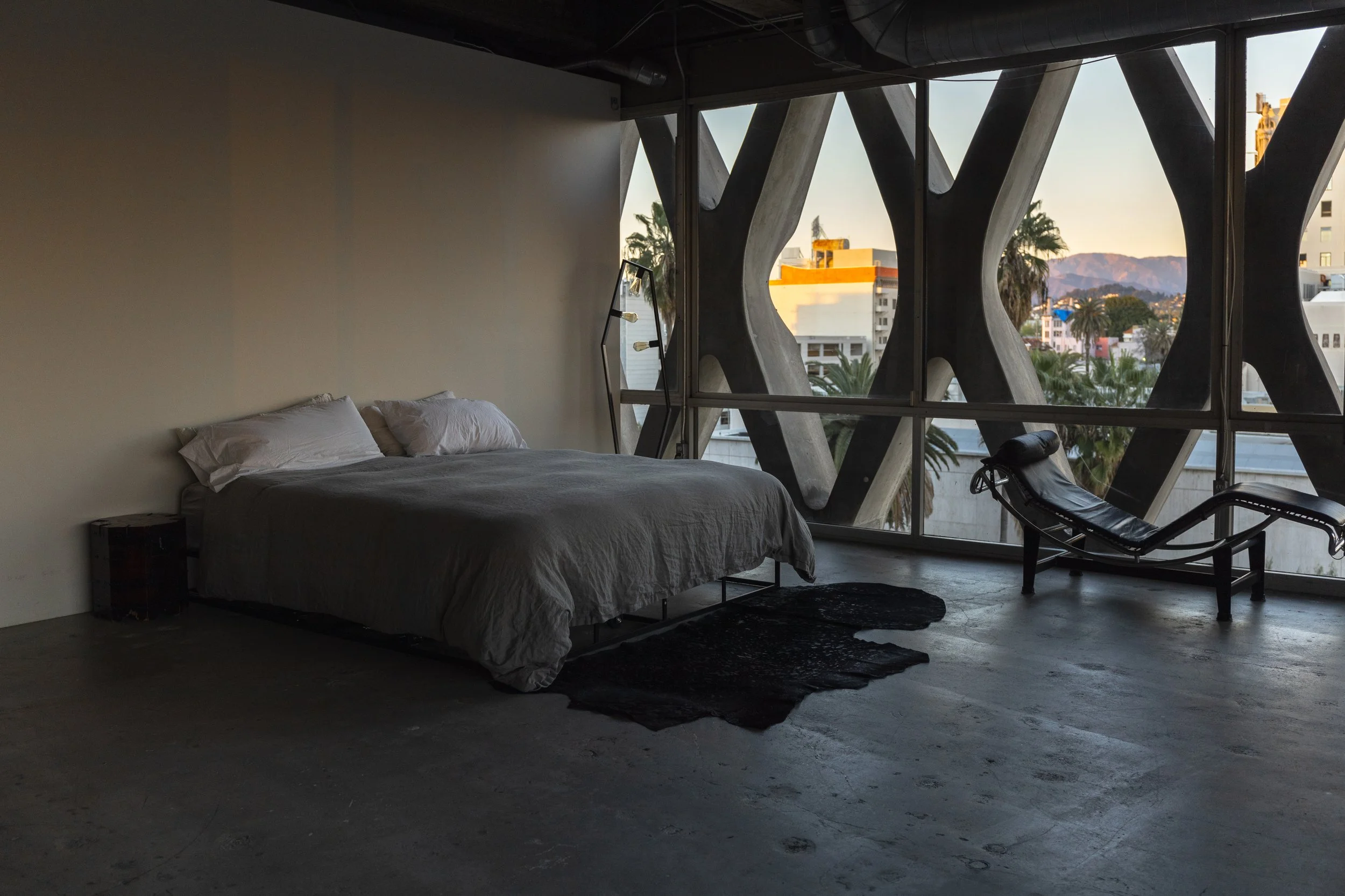 Minimalistic bedroom with a bed, a unique floor lamp, and a black lounge chair near a large window with an urban view and unique architectural supports.