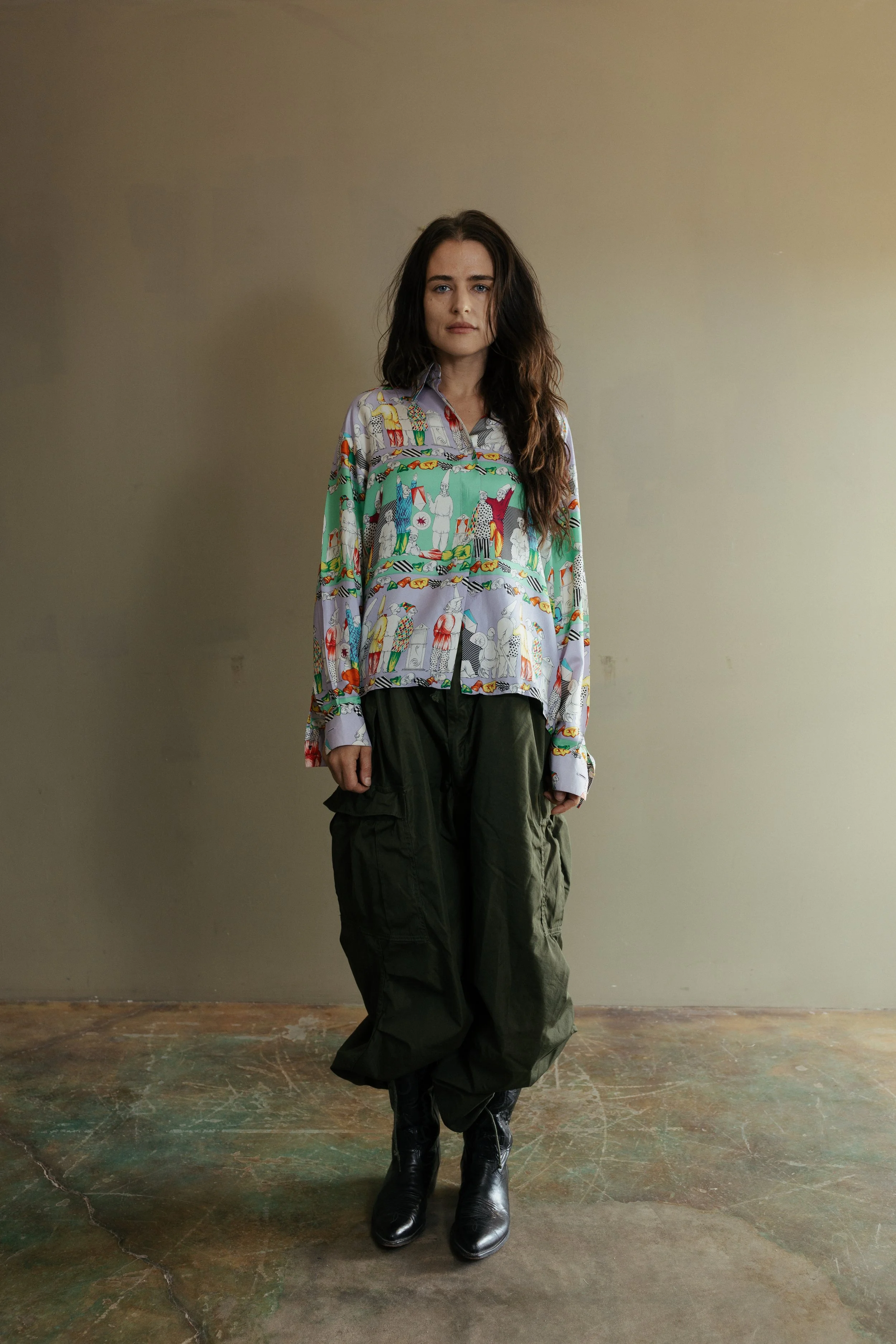 A woman with long, wavy brown hair standing against a plain beige wall, wearing an oversized, colorful shirt with an intricate pattern, dark green cargo pants, and black boots.