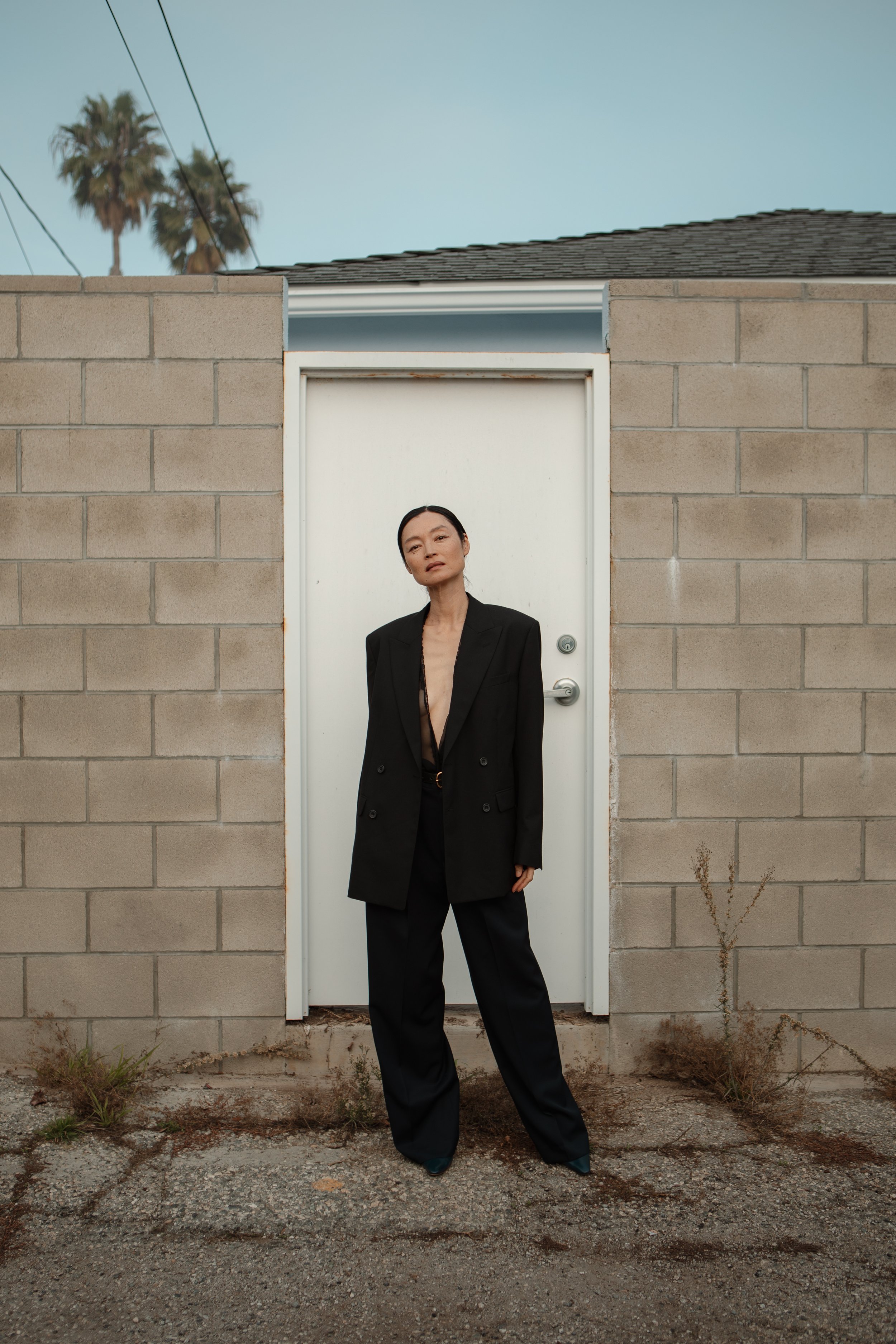 A woman in a black blazer and wide-leg pants standing in front of a white door set into a gray brick wall, with palm trees and power lines in the background.