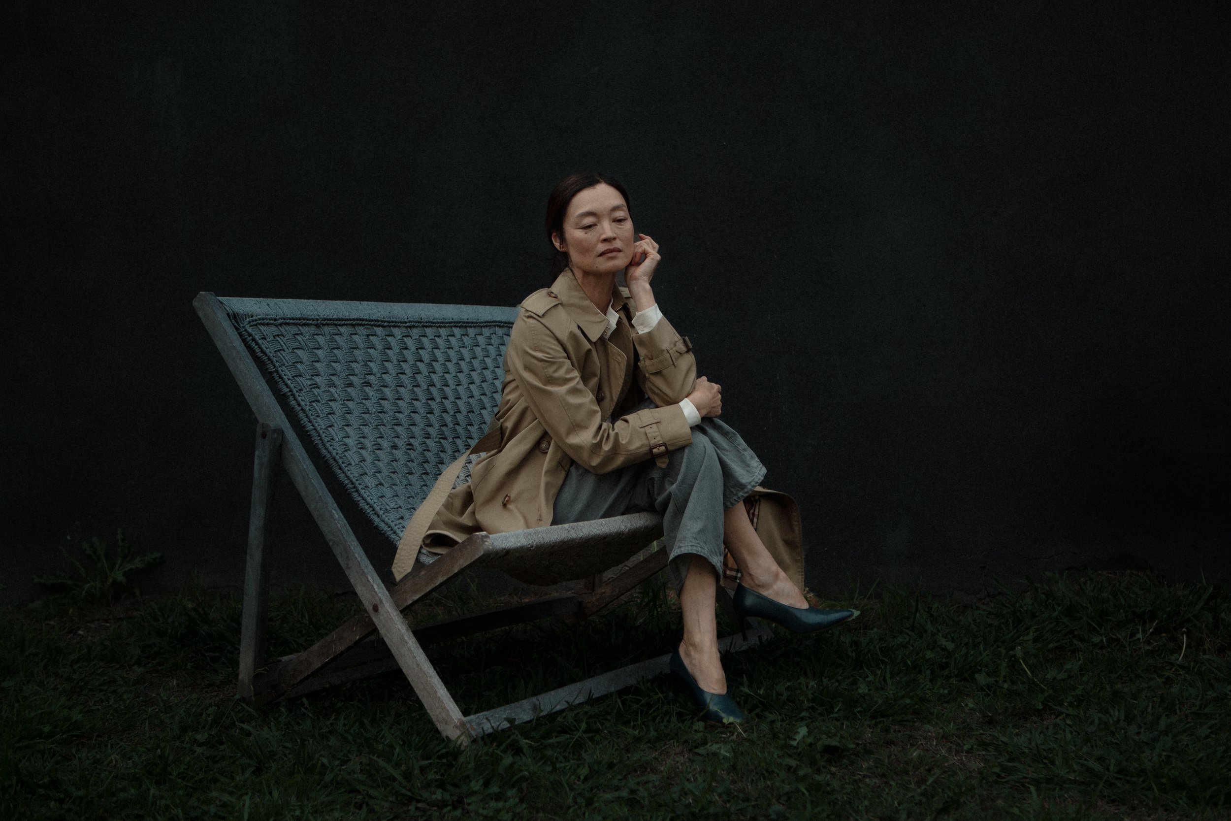 A woman in a beige trench coat, gray pants, and black heels sitting on a wooden lounge chair outdoors against a dark background.