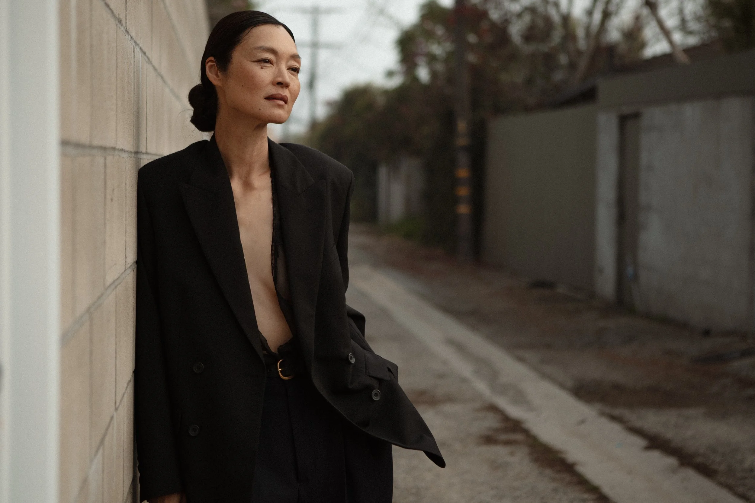 A woman with dark hair tied back, wearing a black blazer with no shirt underneath, leaning against a brick wall in an alleyway during dusk.