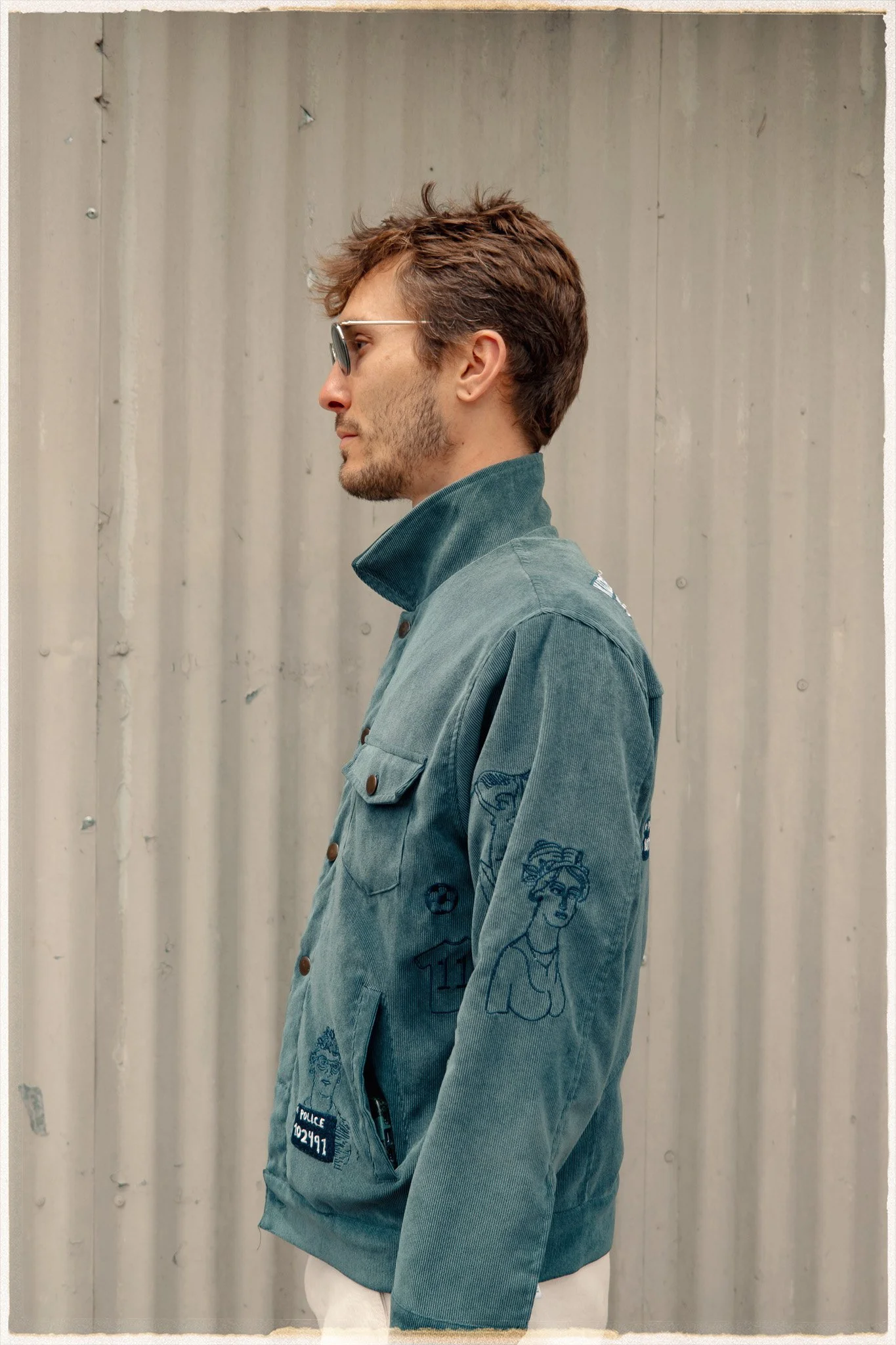 Side profile of a man wearing sunglasses and a denim jacket with multiple patches and embroidered designs, standing against a corrugated metal wall.