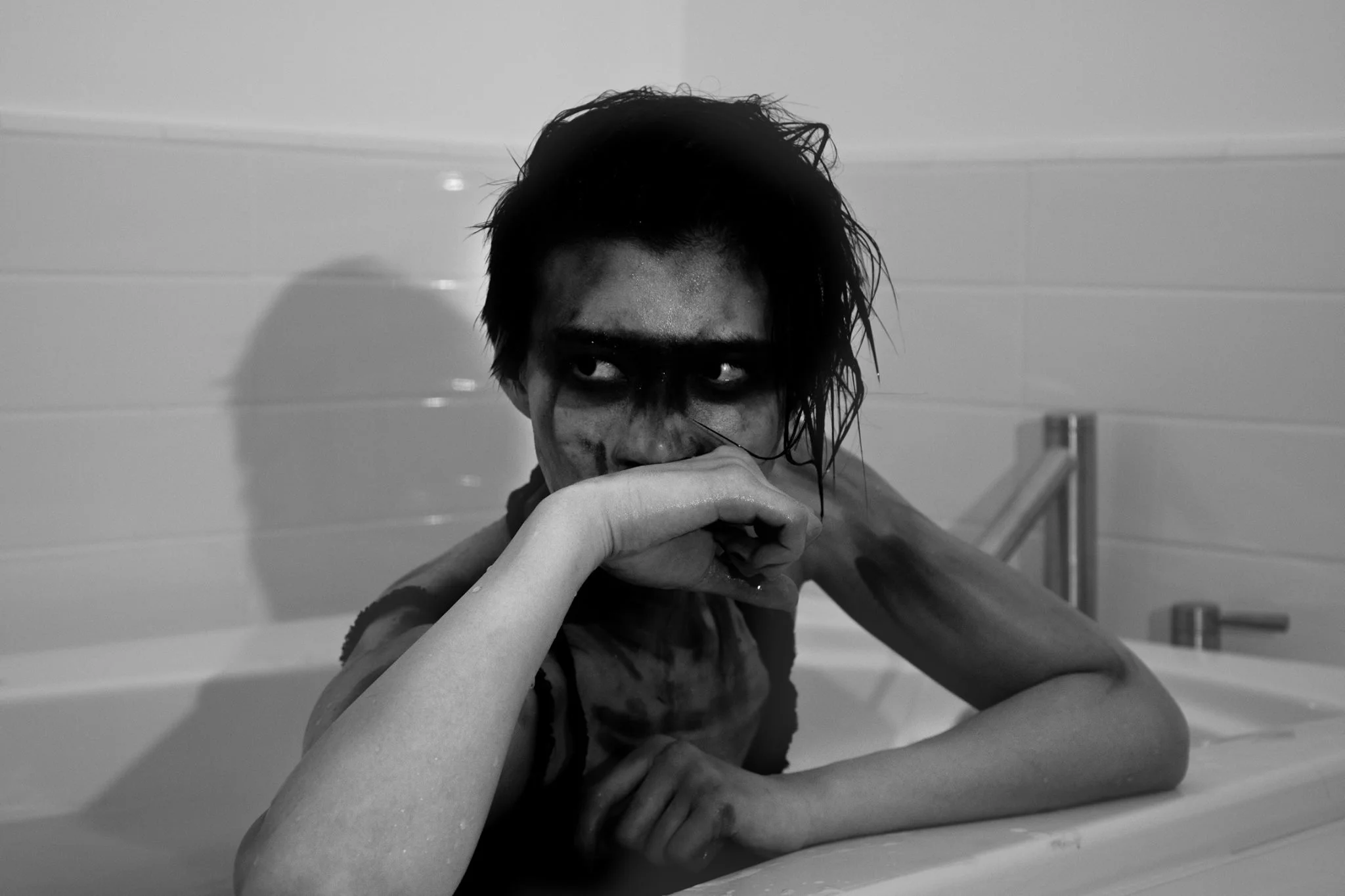 A person with dark, wet hair and dark eye makeup lounging in a bathtub, looking to the side with an intense expression.