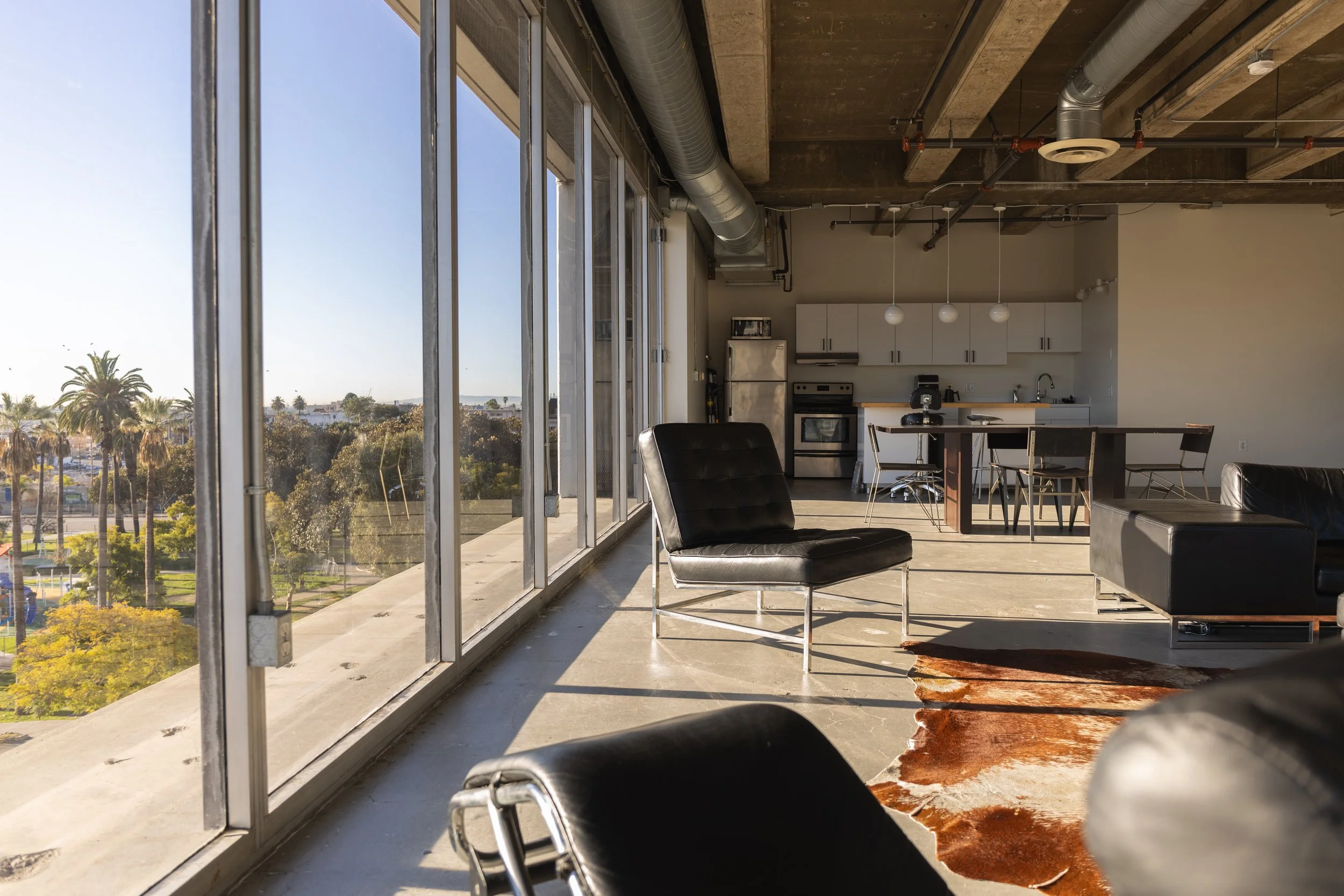 Modern open-concept apartment with large floor-to-ceiling windows, concrete floors, minimalist black furniture, and a kitchen area with white cabinets and wooden island, overlooking palm trees and cityscape outside.