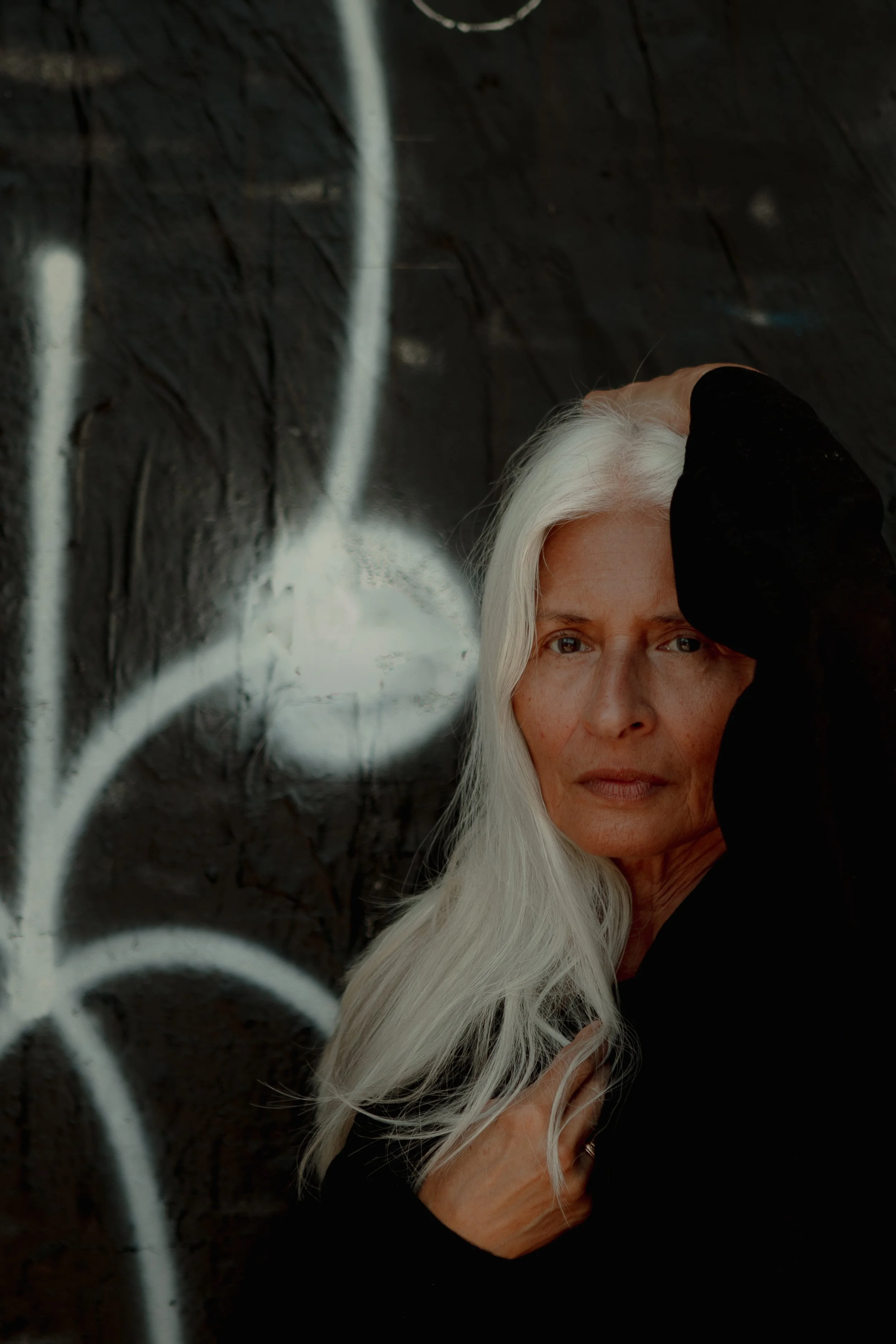 A mature woman with long white hair, wearing a black top, posing against a dark wall with white graffiti.