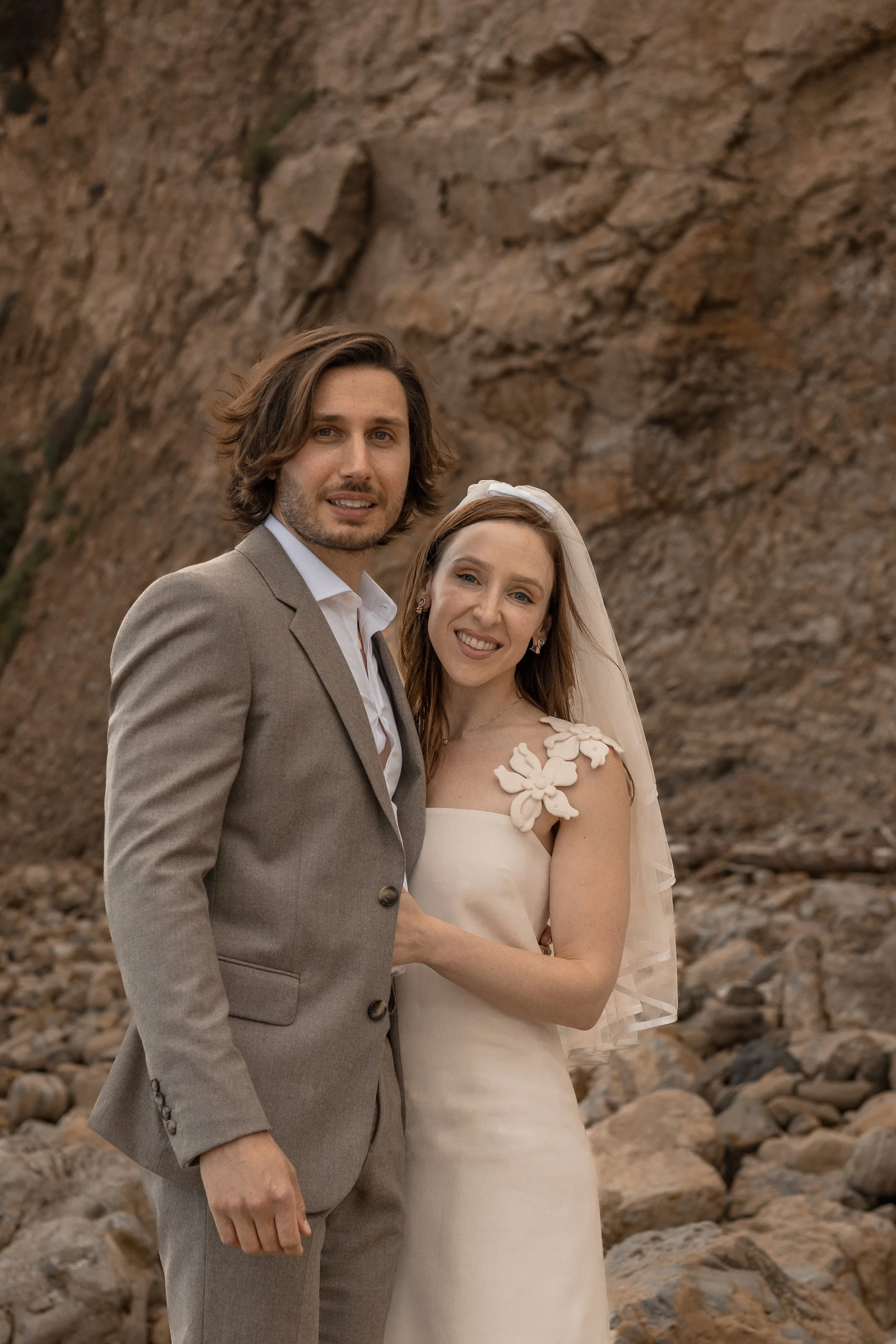 A bride and groom standing together outdoors in front of a rocky cliff, smiling. The groom has shoulder-length brown hair and is wearing a light gray suit, while the bride has long red hair, a veil, and a white dress with floral details on the should