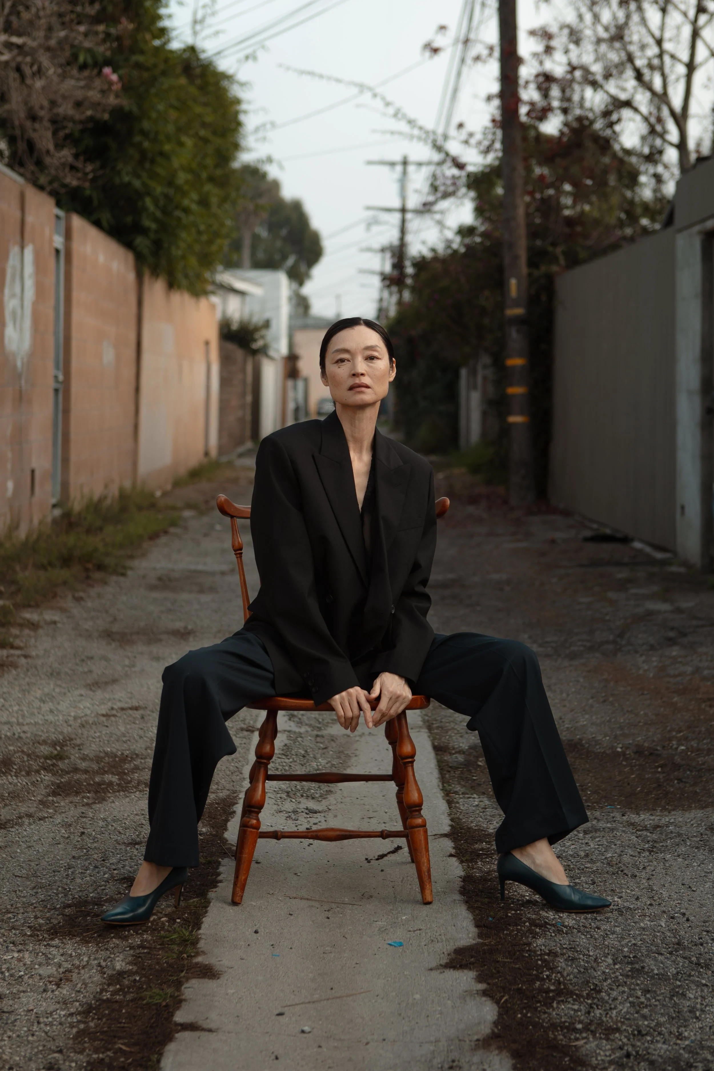 A woman in a black suit with wide leg pants and high heels, sitting on a wooden chair in the middle of an alley with gravel and patches of concrete, flanked by fences and trees, under an overcast sky.