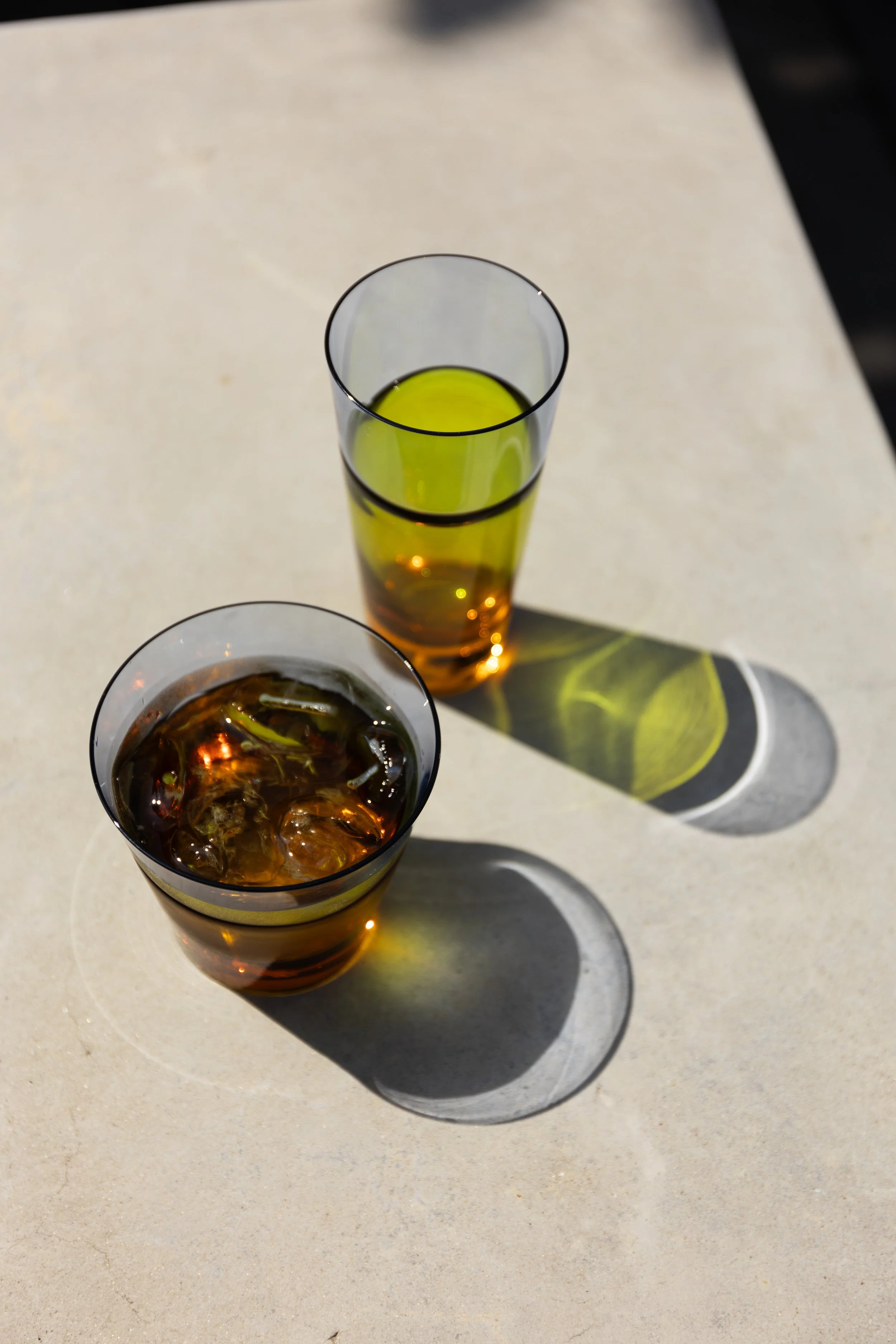 Two glasses with dark and light colored beverages on a light surface, casting distinct shadows.