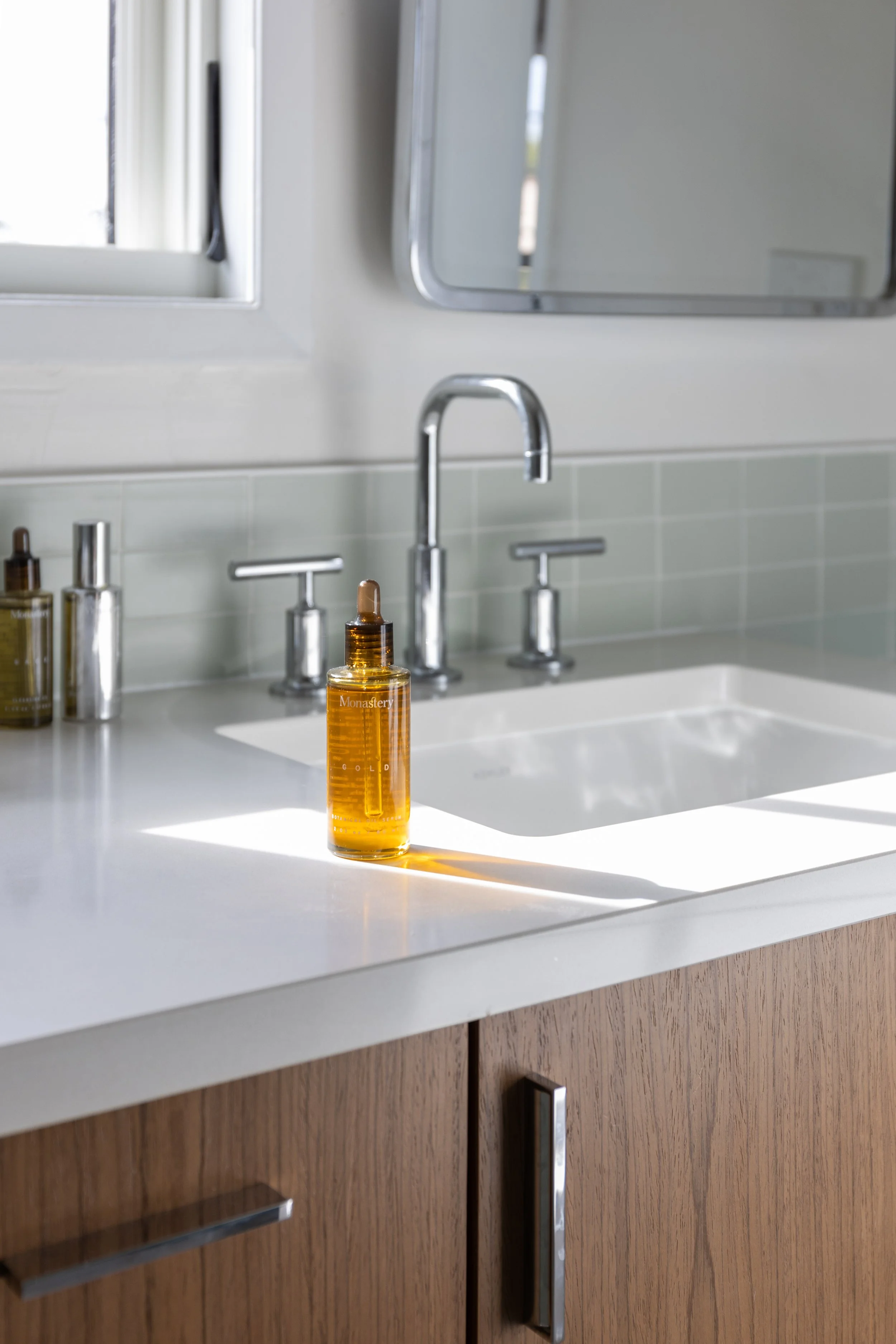 A bathroom sink with a soap or oil dropper bottle labeled Monastery sitting on the white countertop, sunlight casting a shadow, a mirror, and wooden cabinet below.