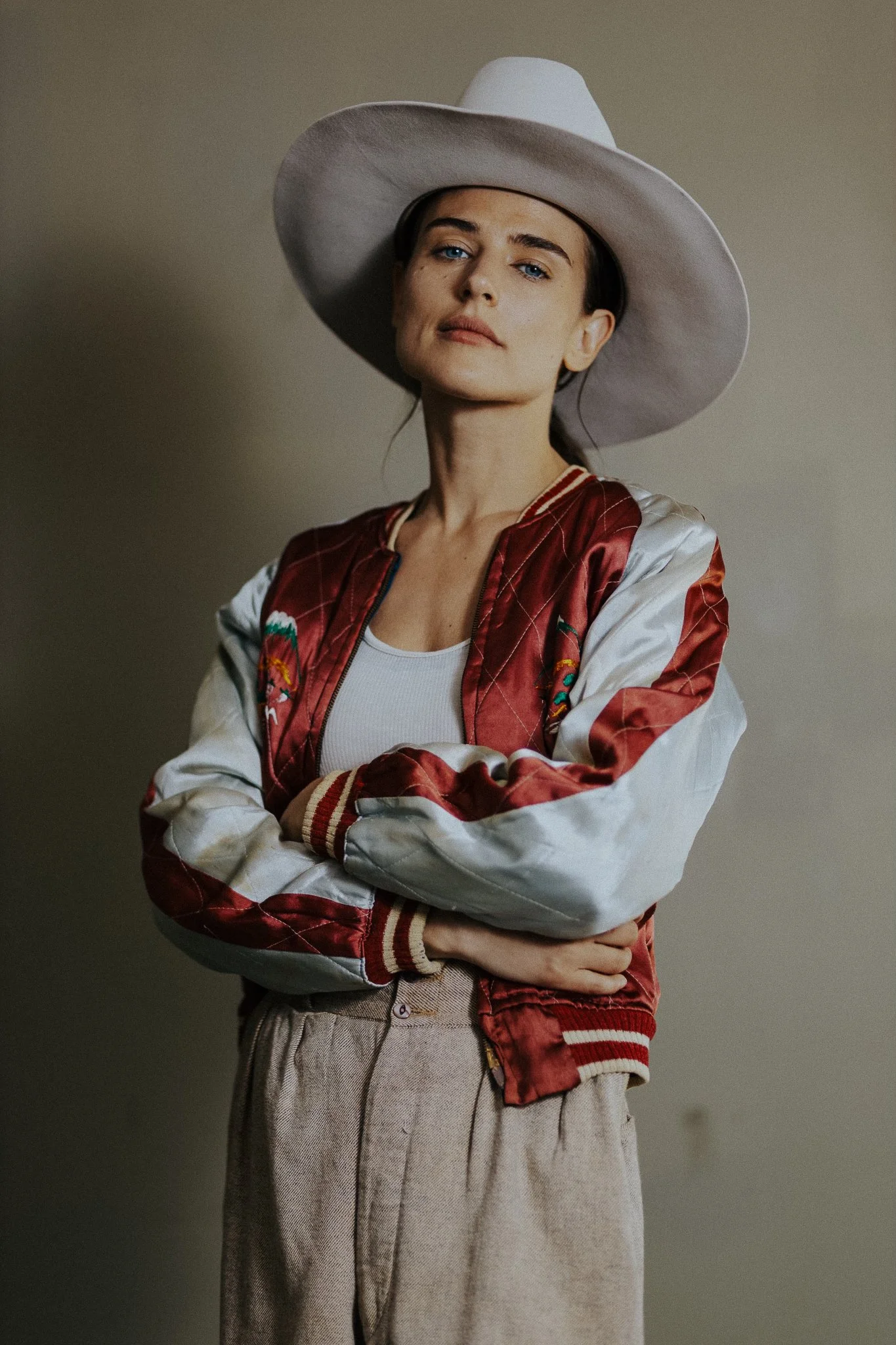 A woman wearing a large white hat, a red and white satin bomber jacket with embroidery, a white shirt, and beige high-waisted pants, standing with arms crossed against a plain background.