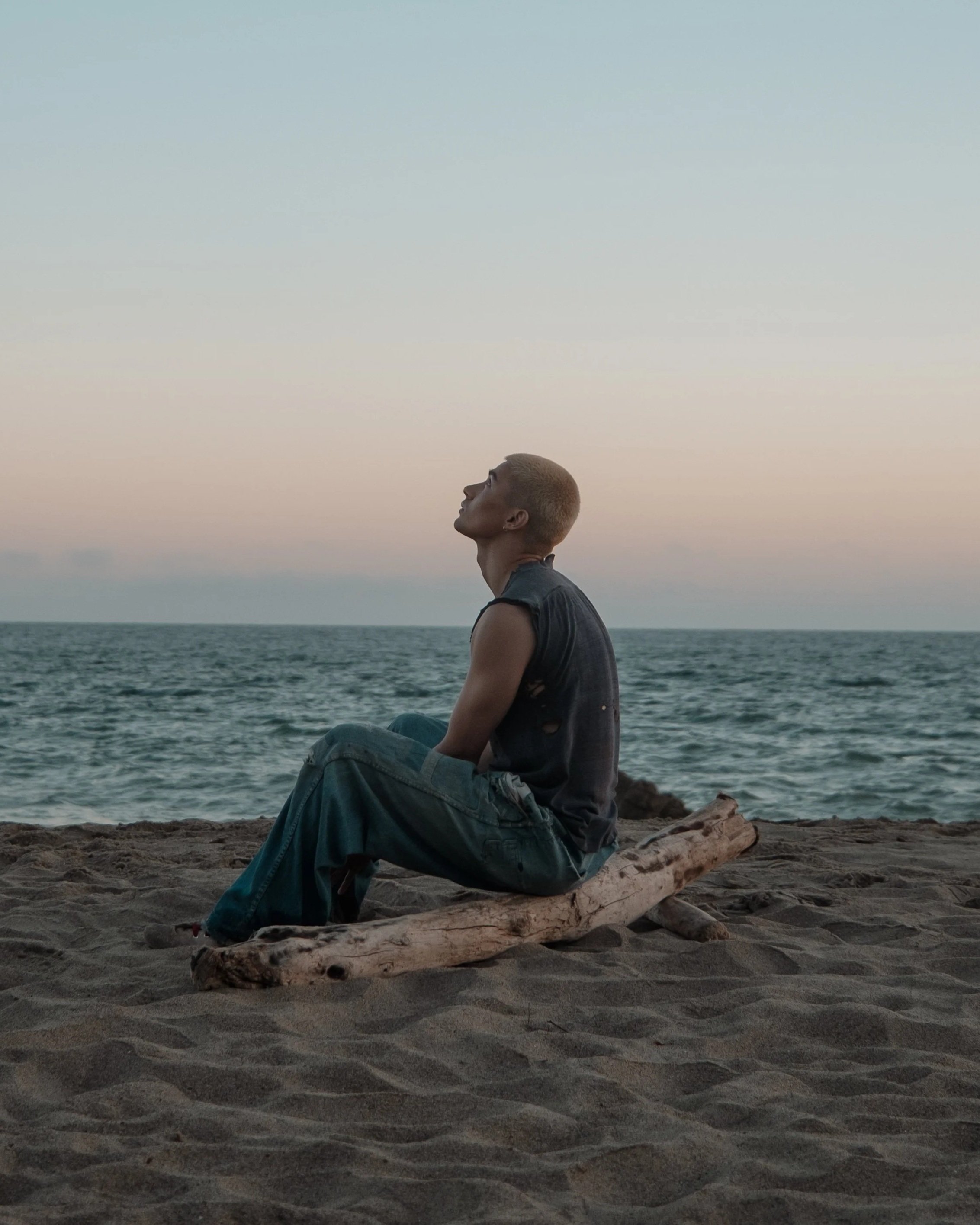 A person with short, blonde hair sitting on a piece of driftwood on the sandy beach, looking up at the sky with the ocean in the background during sunset.