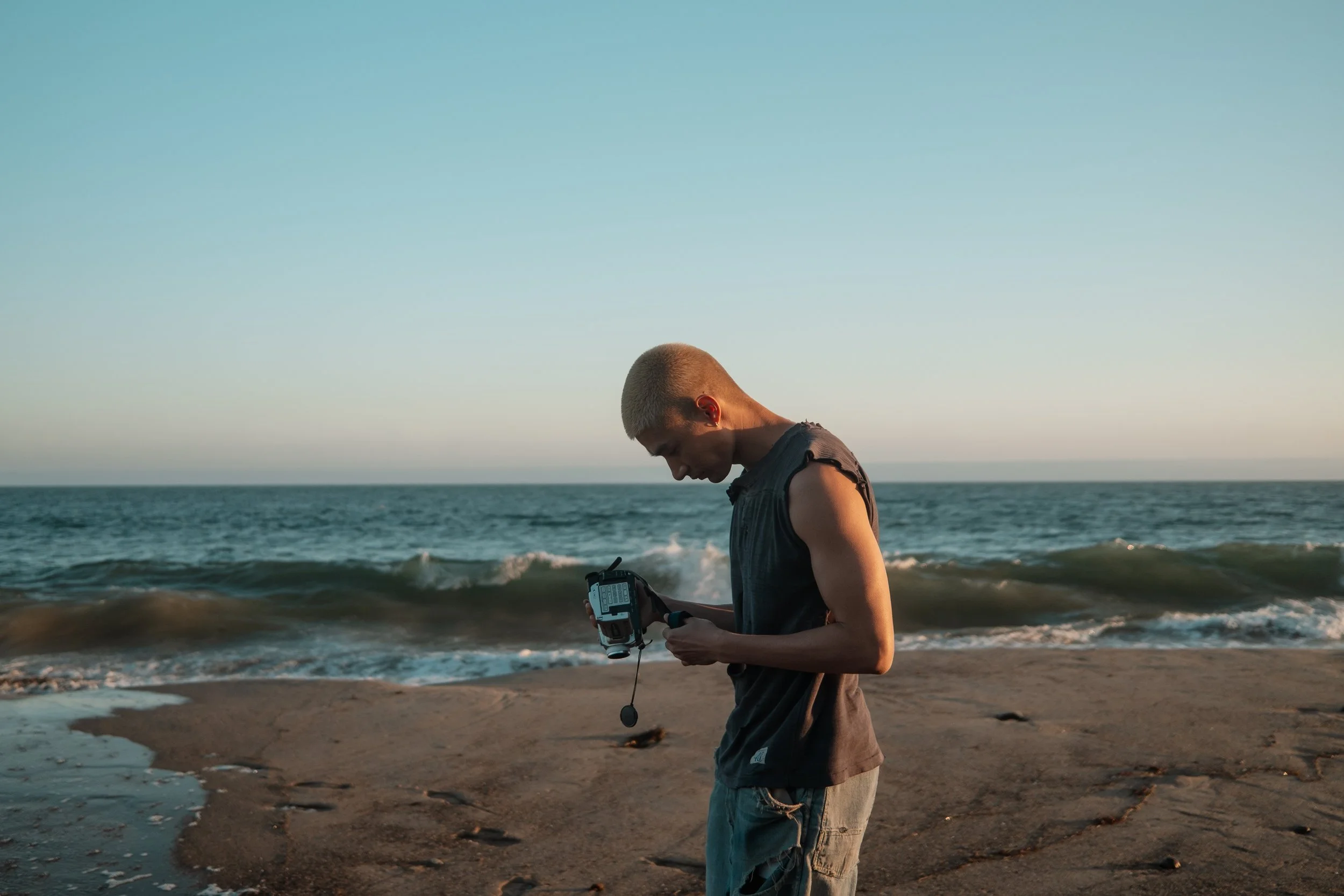 A person with short blonde hair wearing a sleeveless dark shirt and shorts standing on a beach, looking down at a handheld device with water and the ocean in the background.