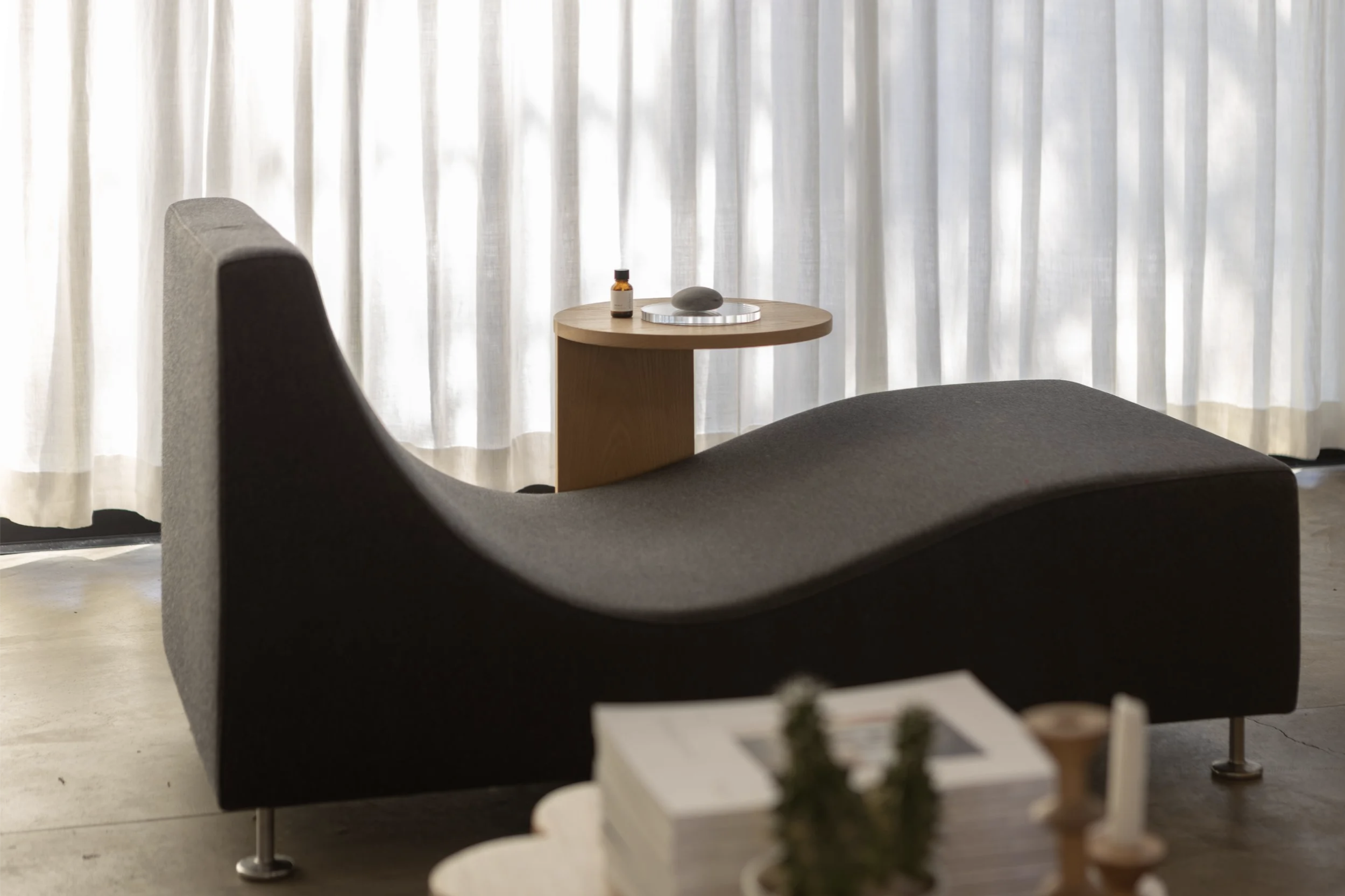 Minimalist living space with black curved lounge chair, wooden side table with small bottle, stone, and tray, white curtains, and a stack of magazines with plants and candles in the foreground.