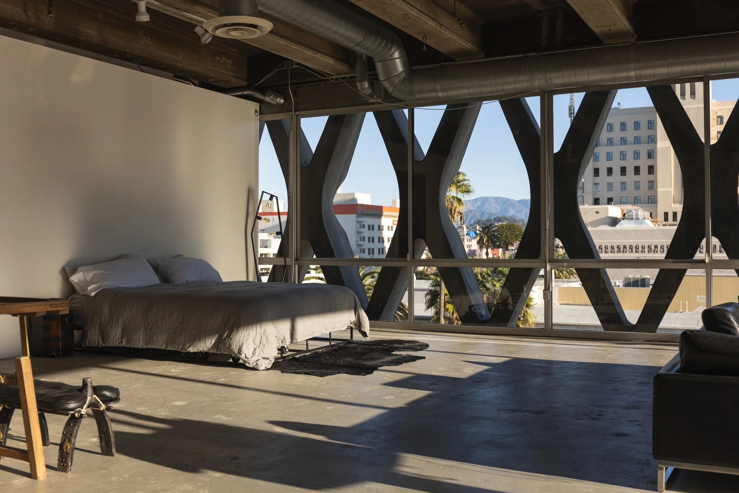 An industrial-style apartment with a bed, a small wooden table, and a chair near a large window with unique architectural supports. City buildings and mountains are visible outside.