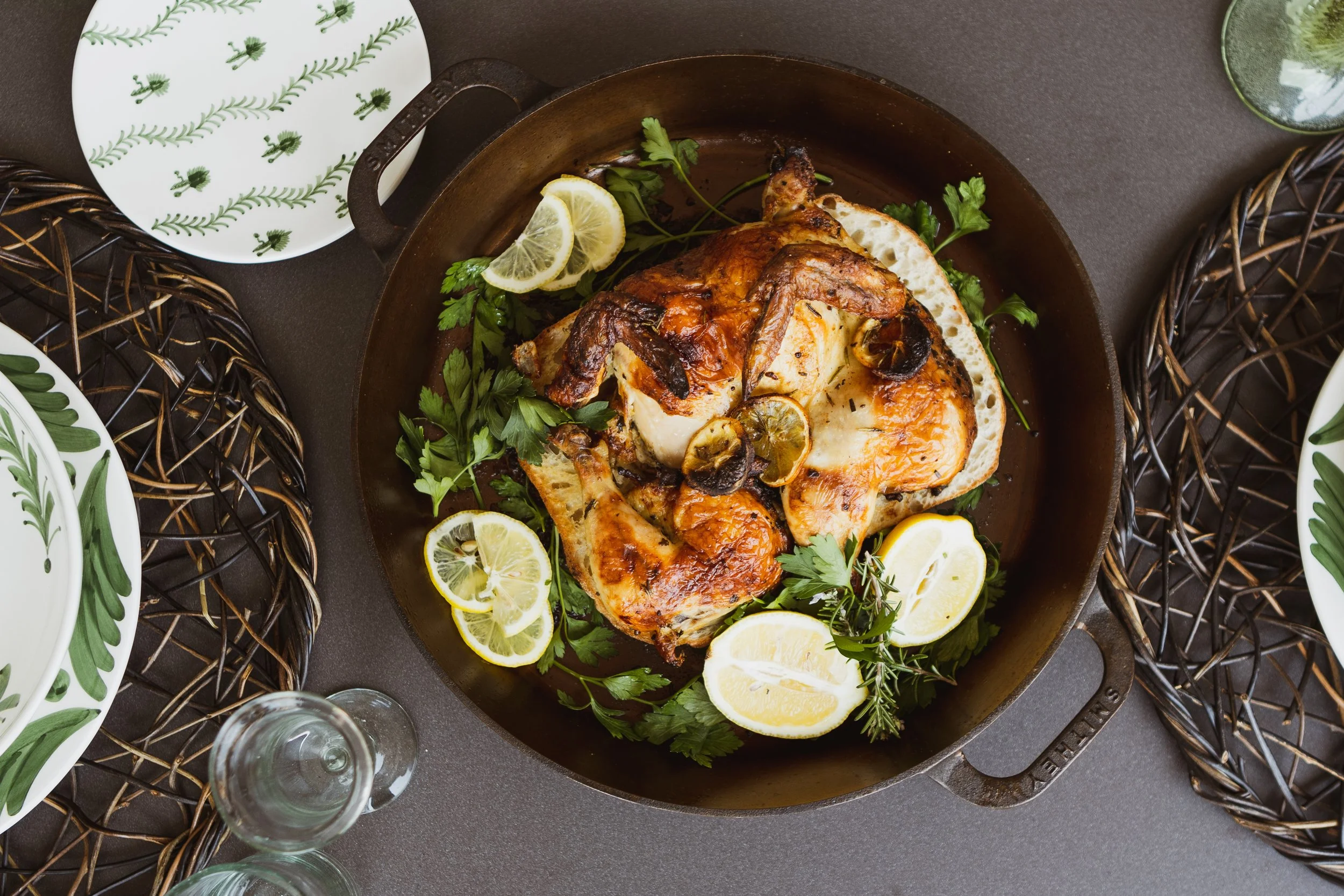 Whole roasted chicken with lemon slices and herbs in a cast iron skillet on a dark table, surrounded by white plates with green leaf patterns and a clear wine glass.
