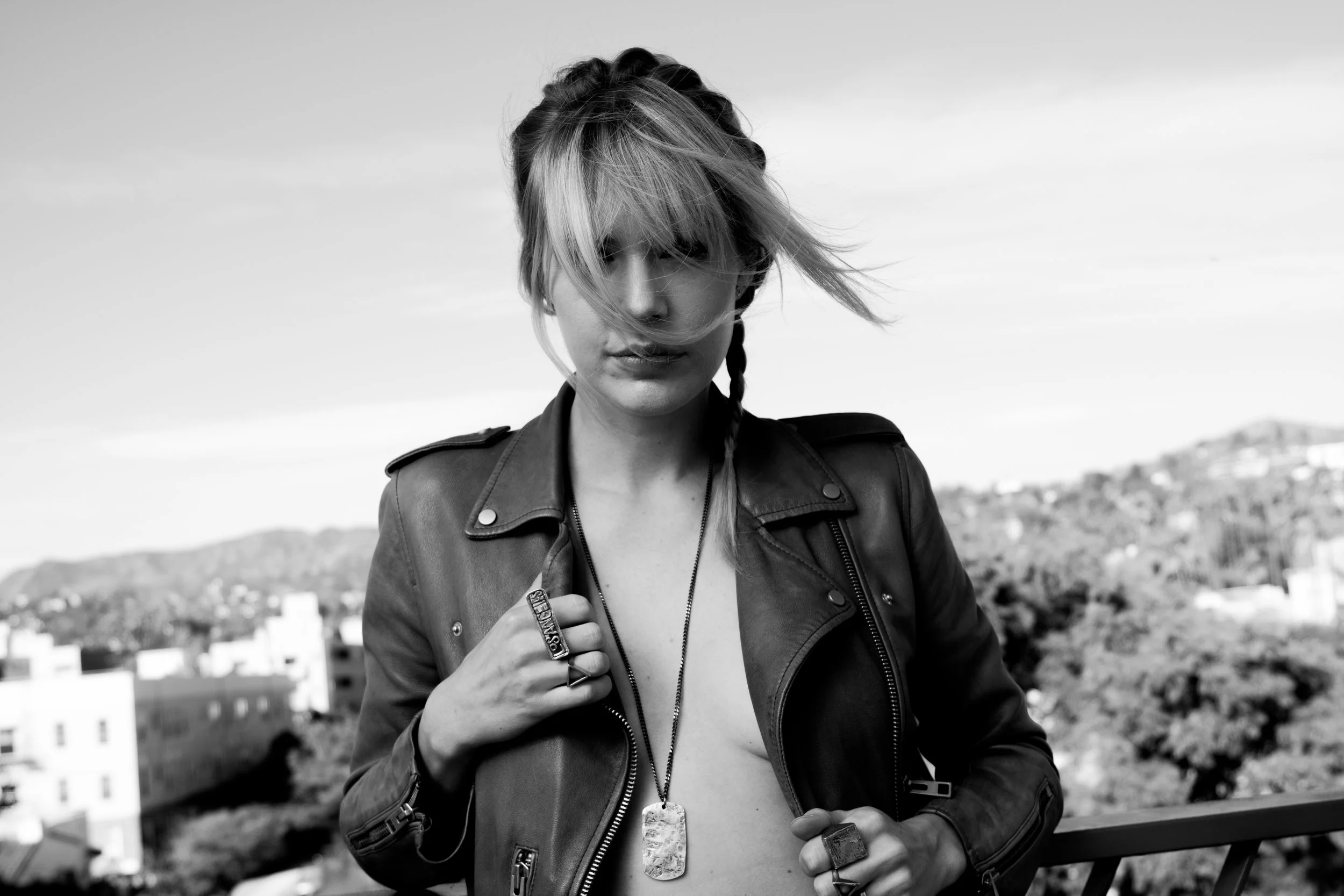 A black and white photo of a woman with shoulder-length hair, wearing a leather jacket, jewelry, and standing outdoors with a cityscape and hills in the background.
