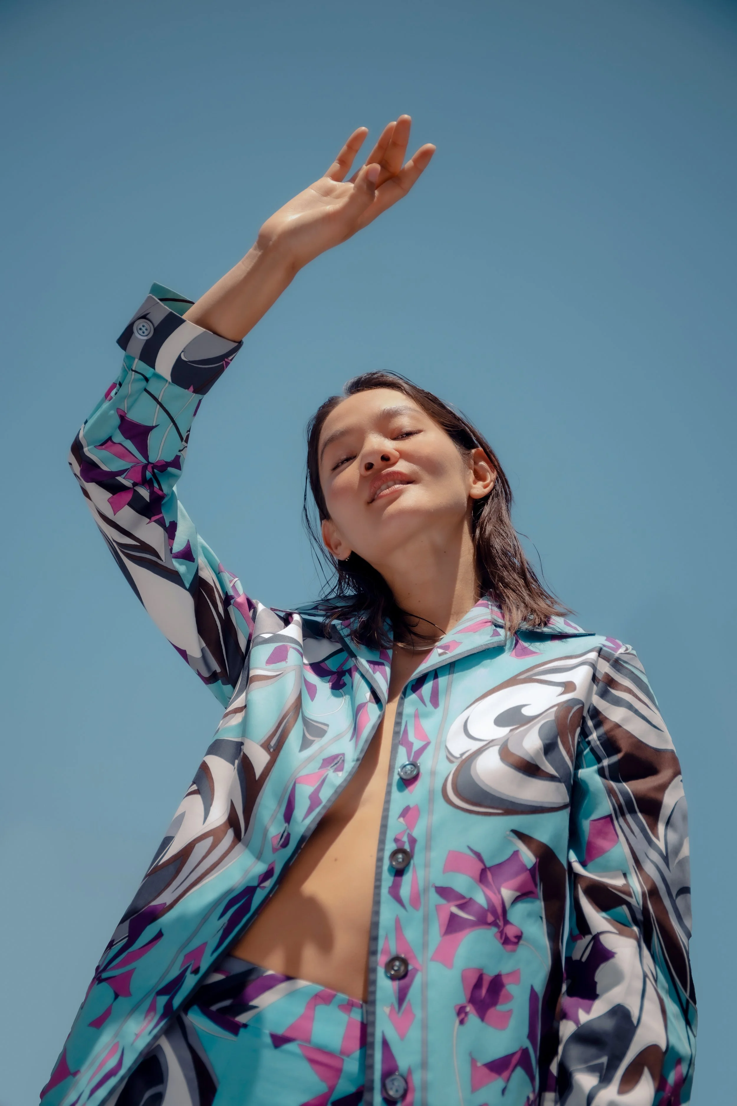 A woman in a colorful patterned jacket and matching pants, standing outdoors with a clear blue sky in the background. She is posing with her arm raised and smiling slightly.
