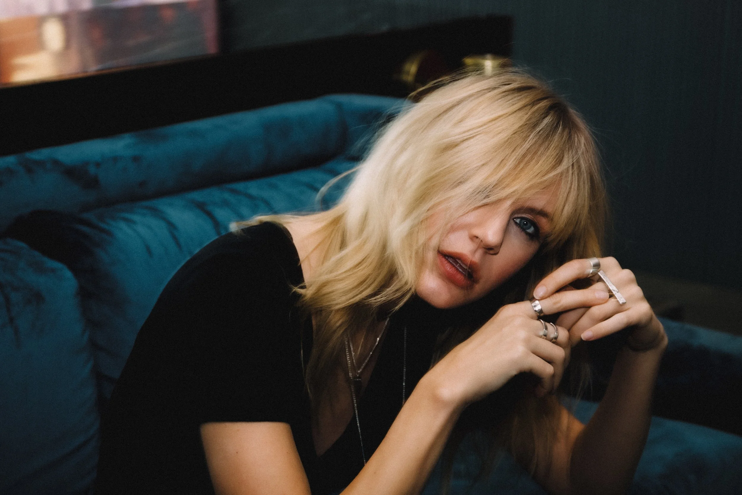 A blonde woman with blue eyes wearing a black top, sitting on a dark teal velvet couch, holding several rings in her hands, with hair partially covering her face and an intense expression.