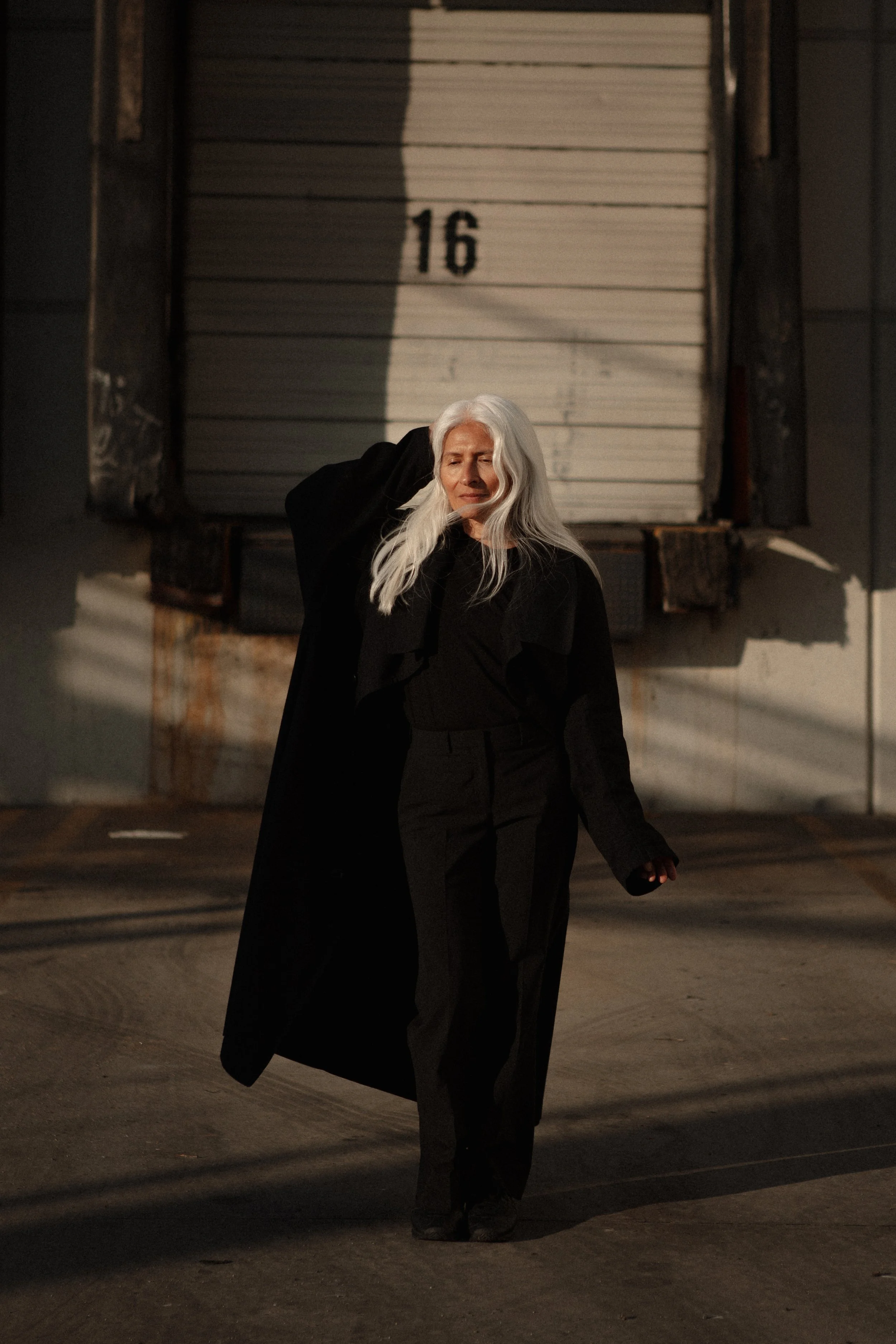 An elderly woman with long white hair walking outdoors at sunset, holding her black coat over her shoulder, with a large closed door numbered 16 in the background.