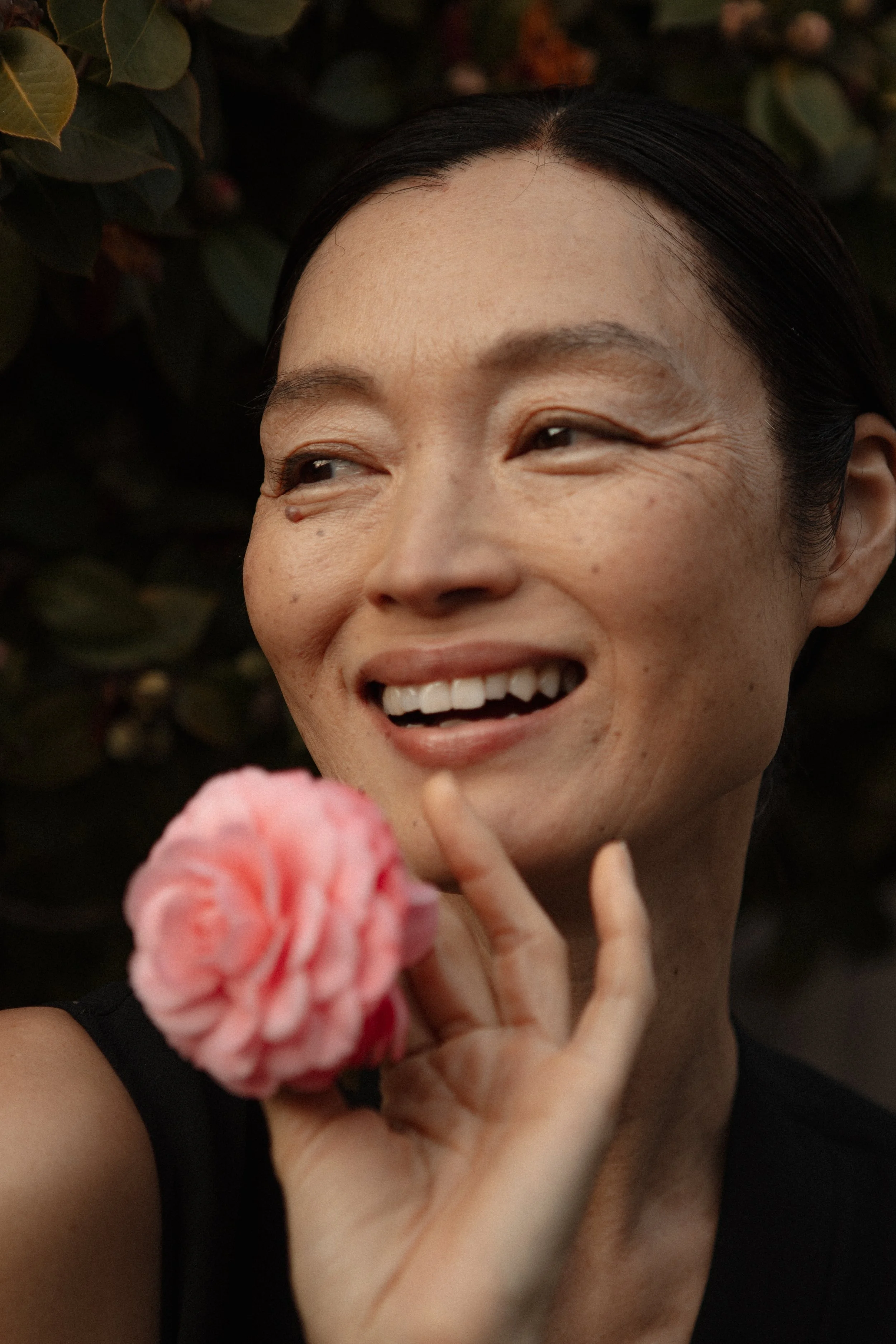 A smiling woman with dark hair holding a pink flower near her face, outdoors with dark green foliage in the background.