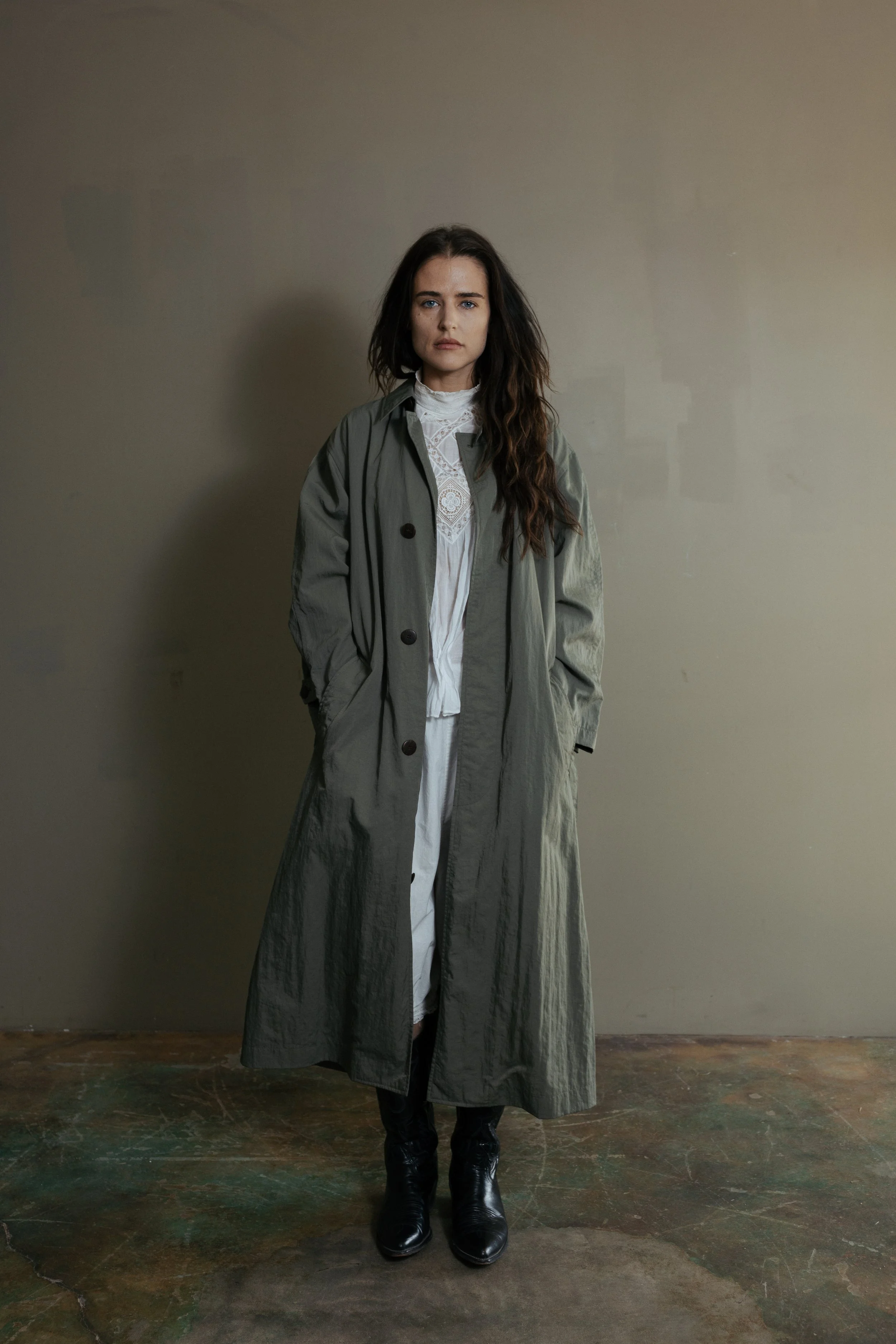 A woman with dark hair standing against a plain wall, wearing a long gray trench coat, a white lace blouse, and black boots.