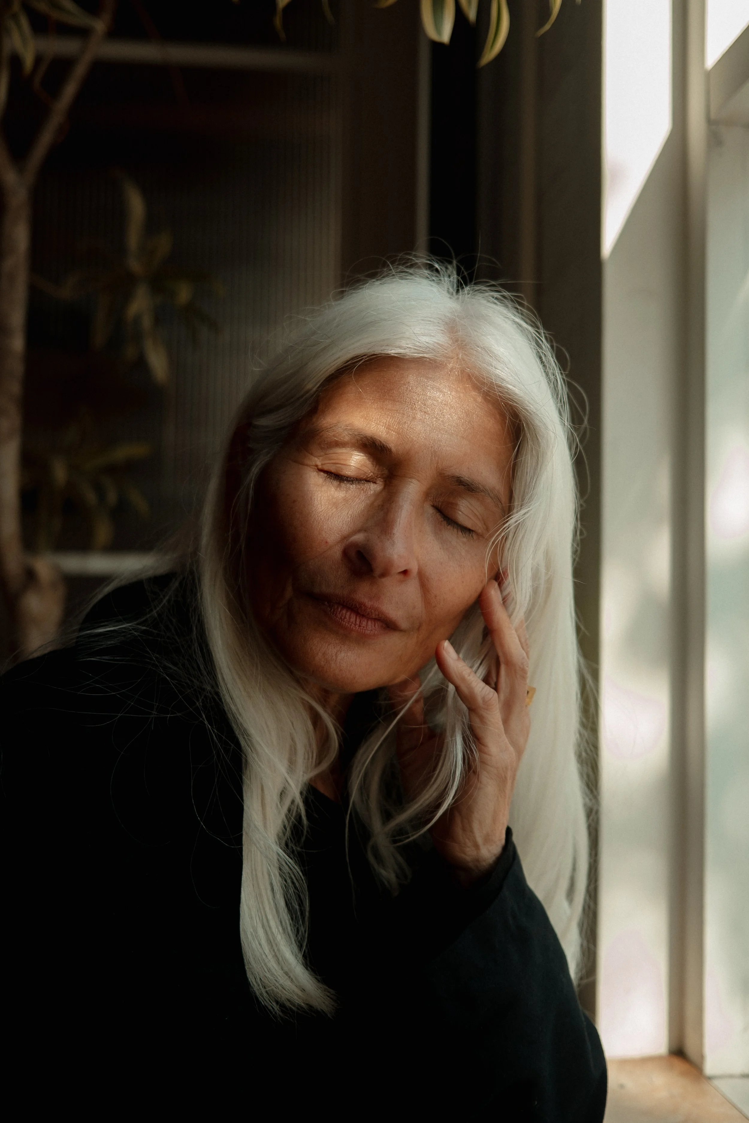 An elderly woman with long white hair resting her head against her hand with eyes closed near a window.