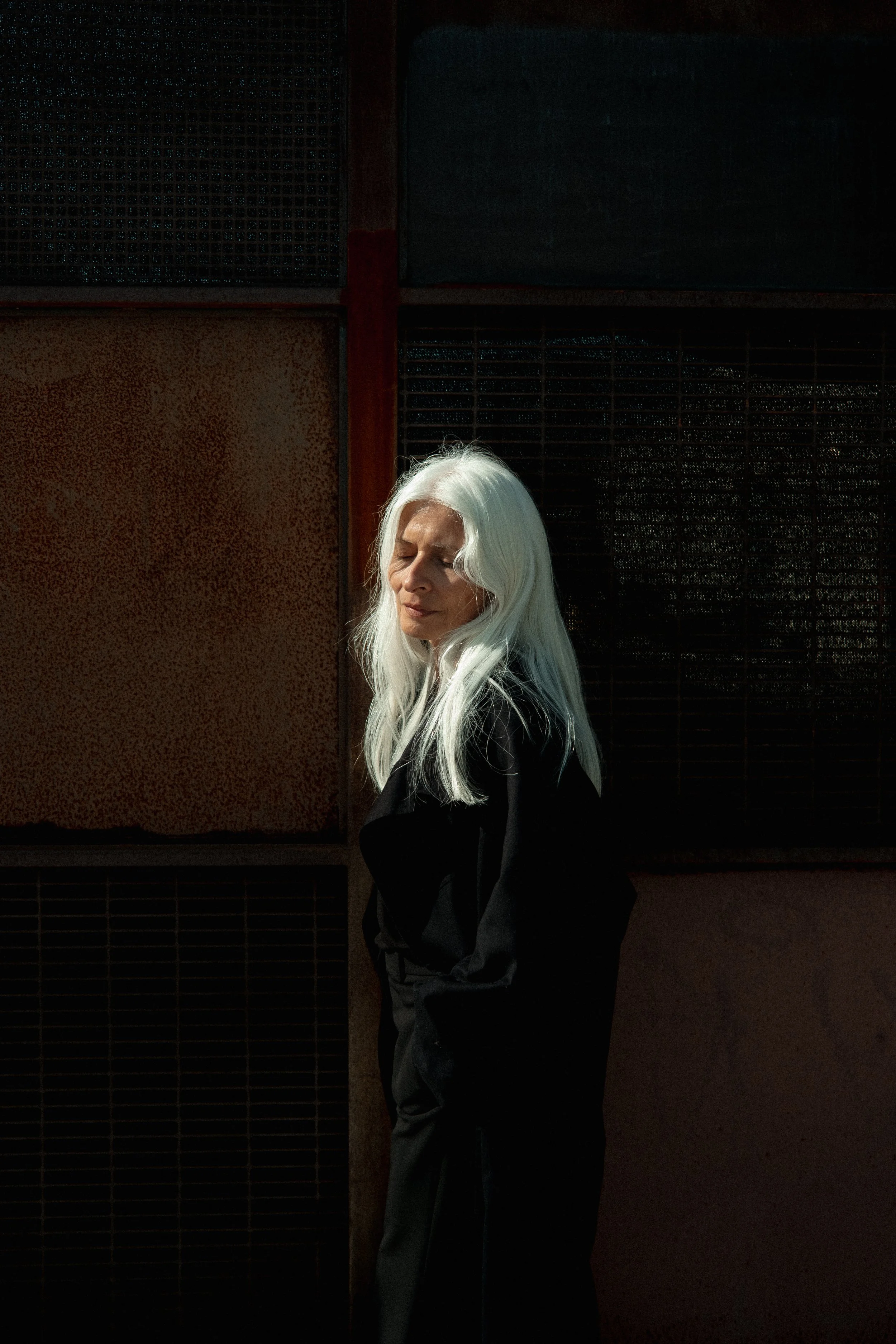An elderly woman with long white hair, dressed in black, standing with her eyes closed against a dark, textured background with various panels.