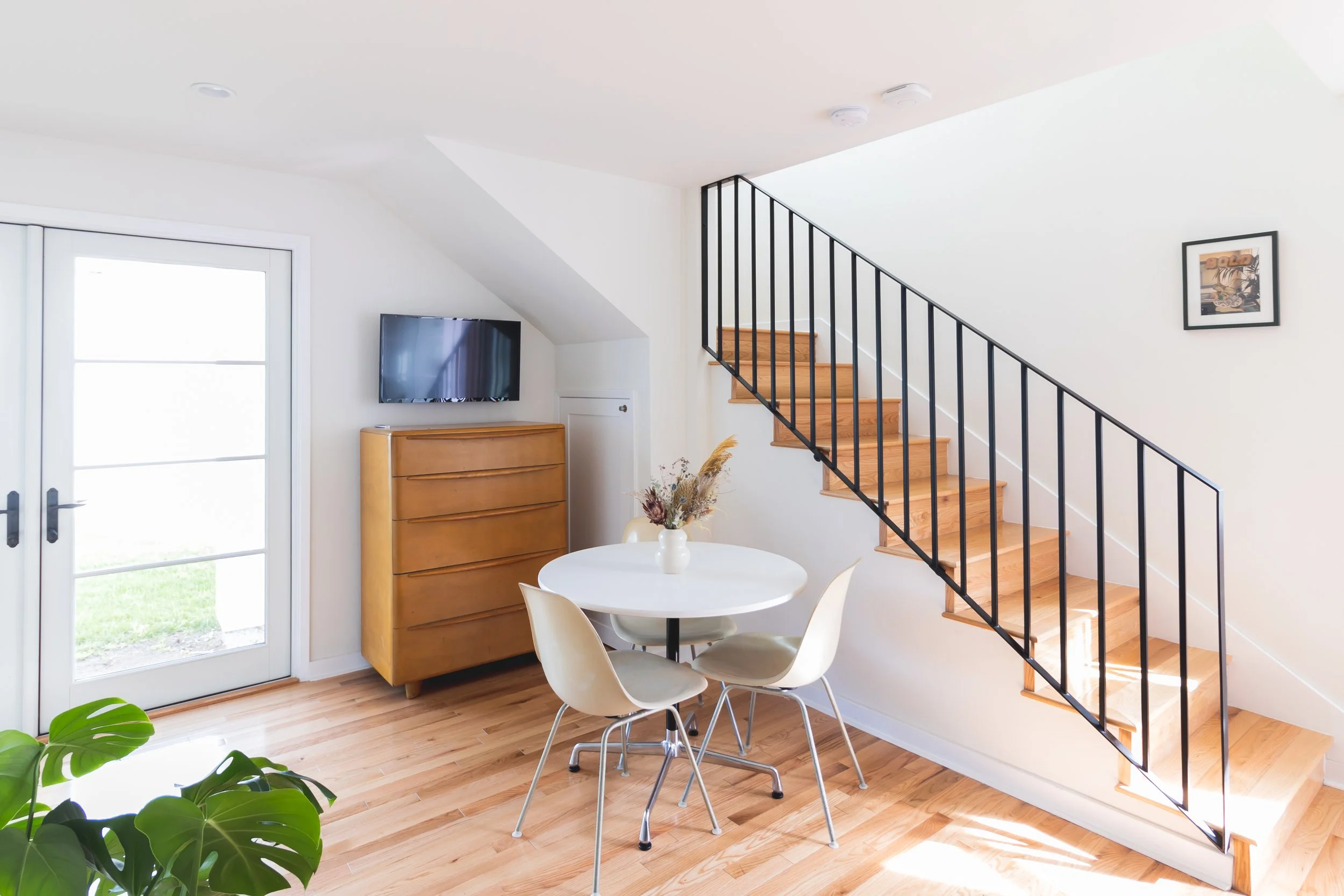 Bright living area with hardwood flooring, a round dining table surrounded by four beige chairs, a small wooden dresser with a TV on top, a staircase with black railing, a framed picture on the wall, and a door leading outside.