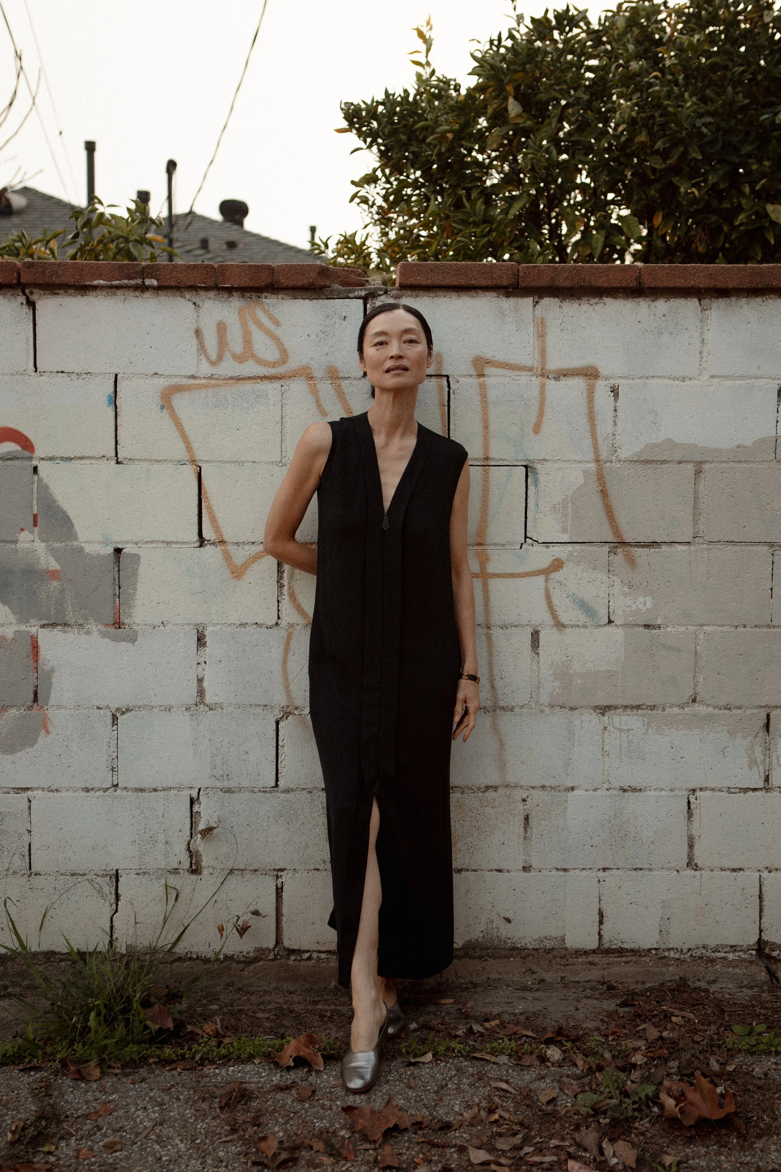A woman in a long black dress and silver heels standing against a graffiti-covered brick wall outdoors at dusk.