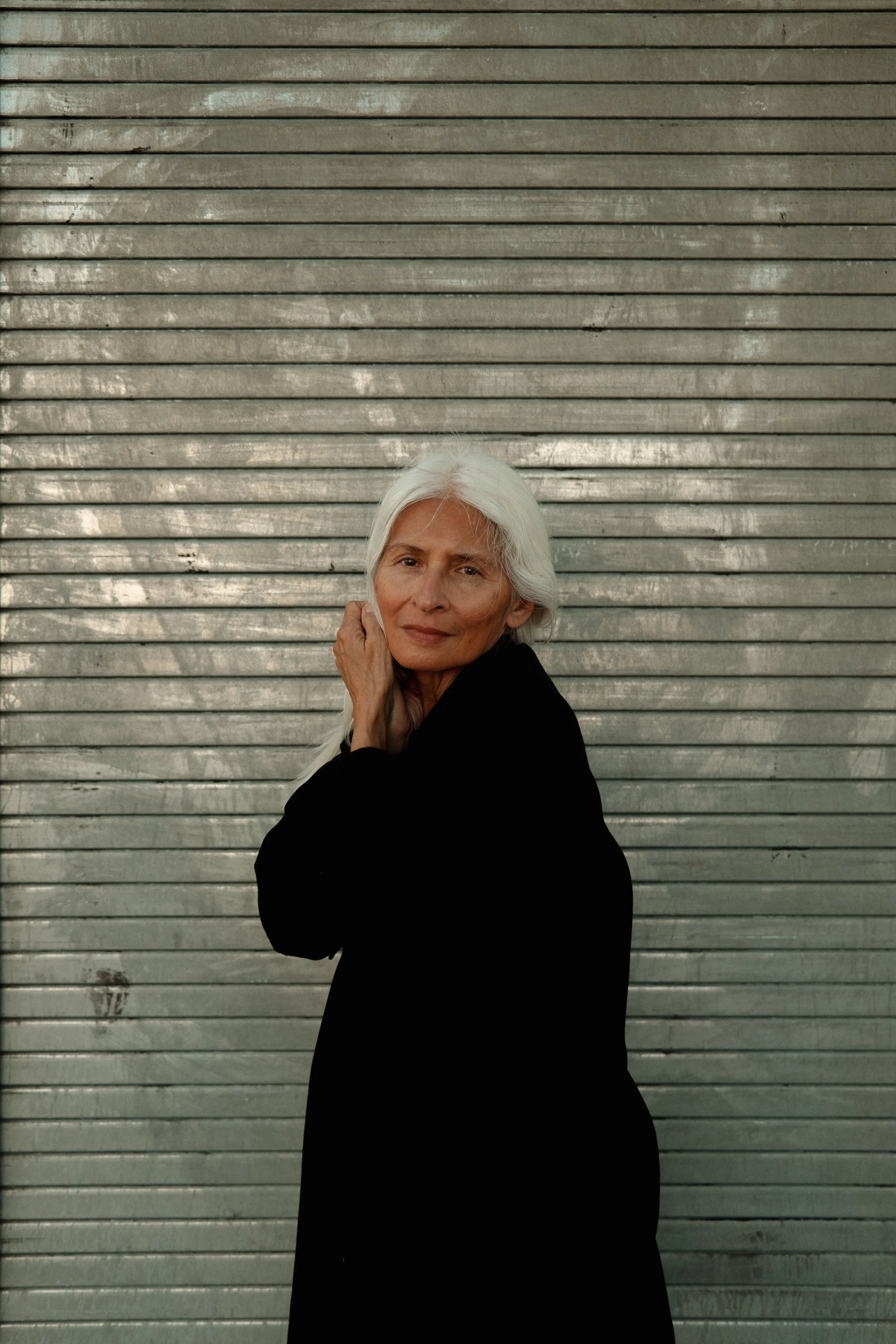 An elderly woman with long white hair, wearing a black coat, standing in front of a metallic garage door with horizontal slats.
