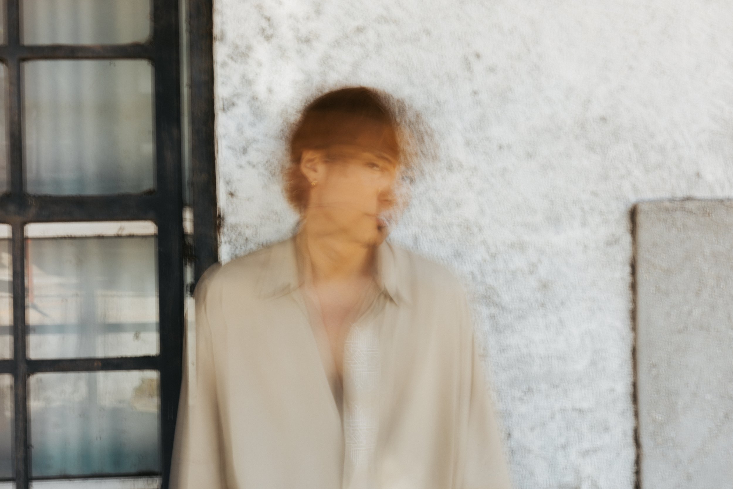 A woman standing near a textured white wall and a window with black metal framing, with her face and motion blurred.