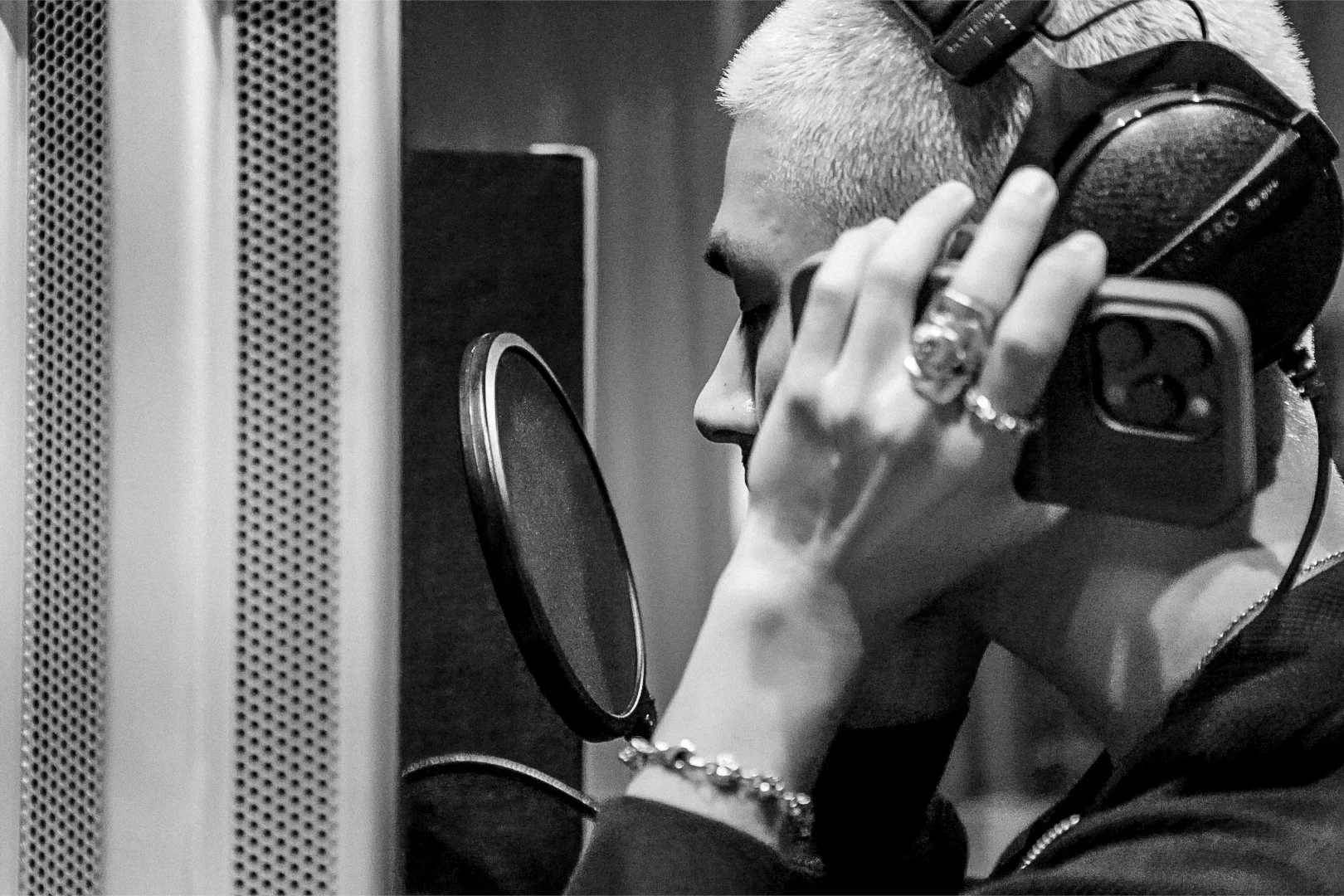 A person with light hair, wearing headphones and multiple rings, is in a recording studio, singing or speaking into a microphone with a pop filter.