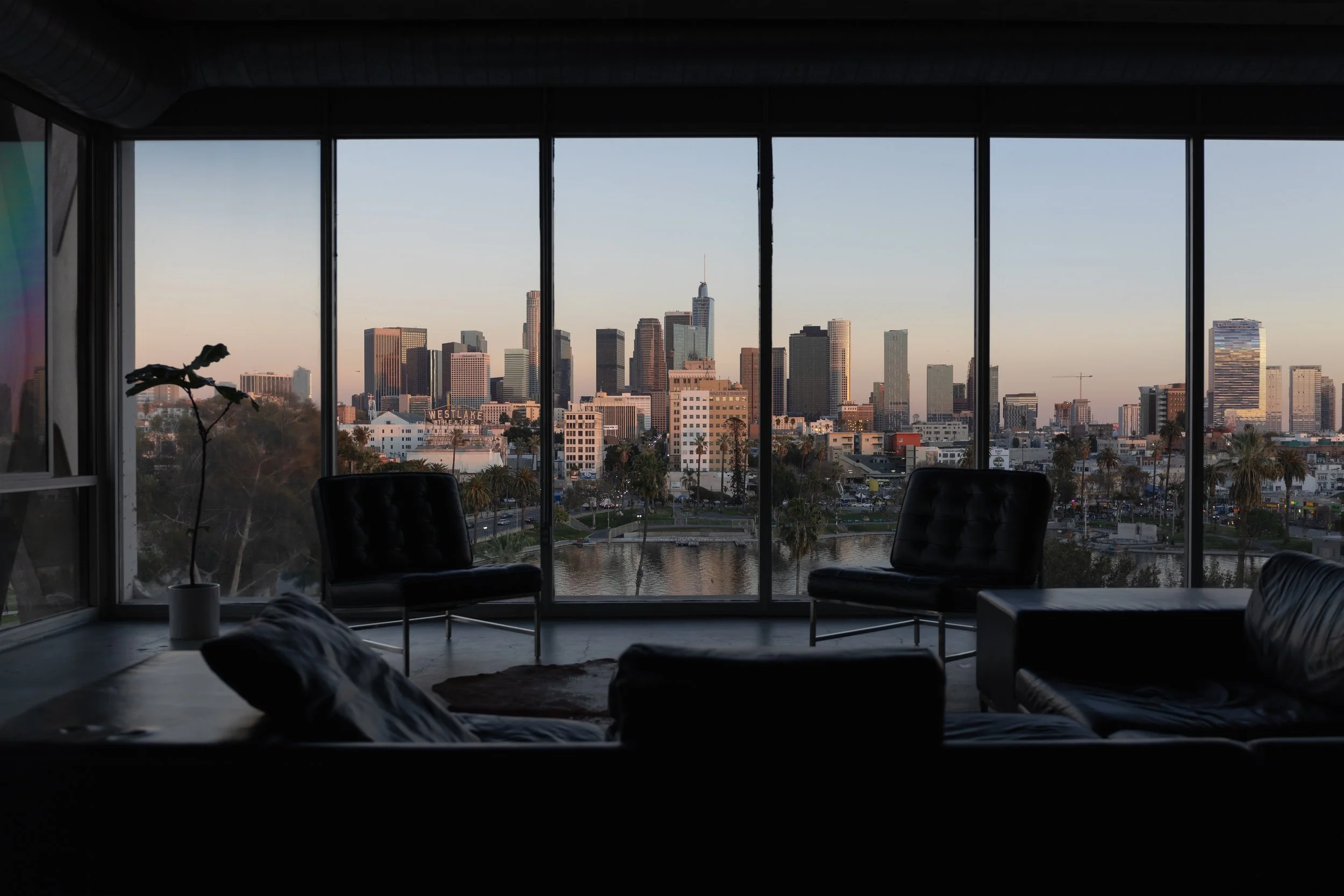 Interior of a modern apartment or hotel room with large floor-to-ceiling windows showcasing a city skyline with tall skyscrapers and buildings, during sunset. The room has black leather chairs and a sofa, and a potted plant is visible on the left sid