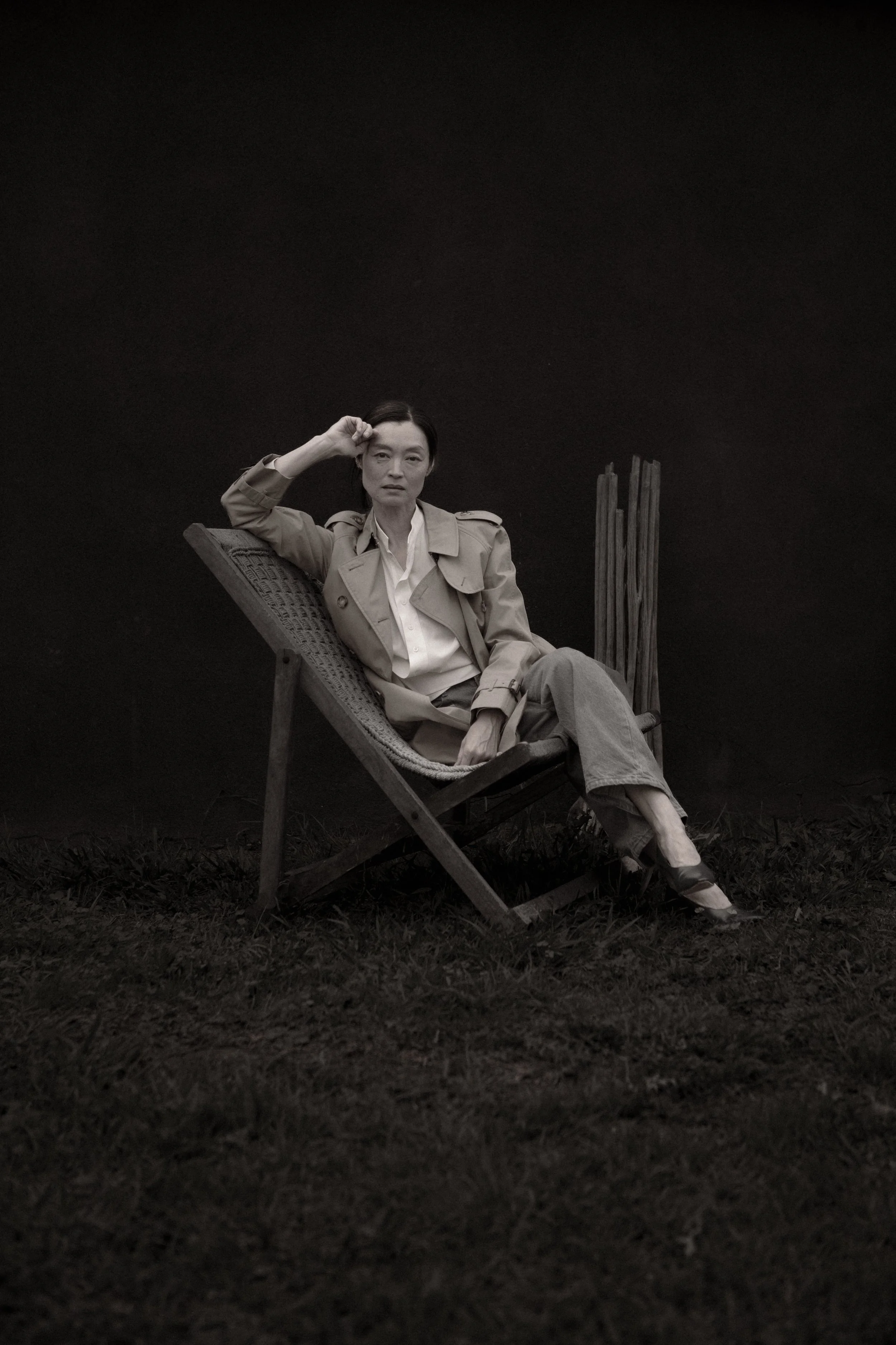 Black-and-white photograph of a woman sitting on a folding chair outdoors against a black background. She is dressed in a trench coat, white shirt, and pants, with one hand resting on her forehead and the other on her lap, looking directly at the cam