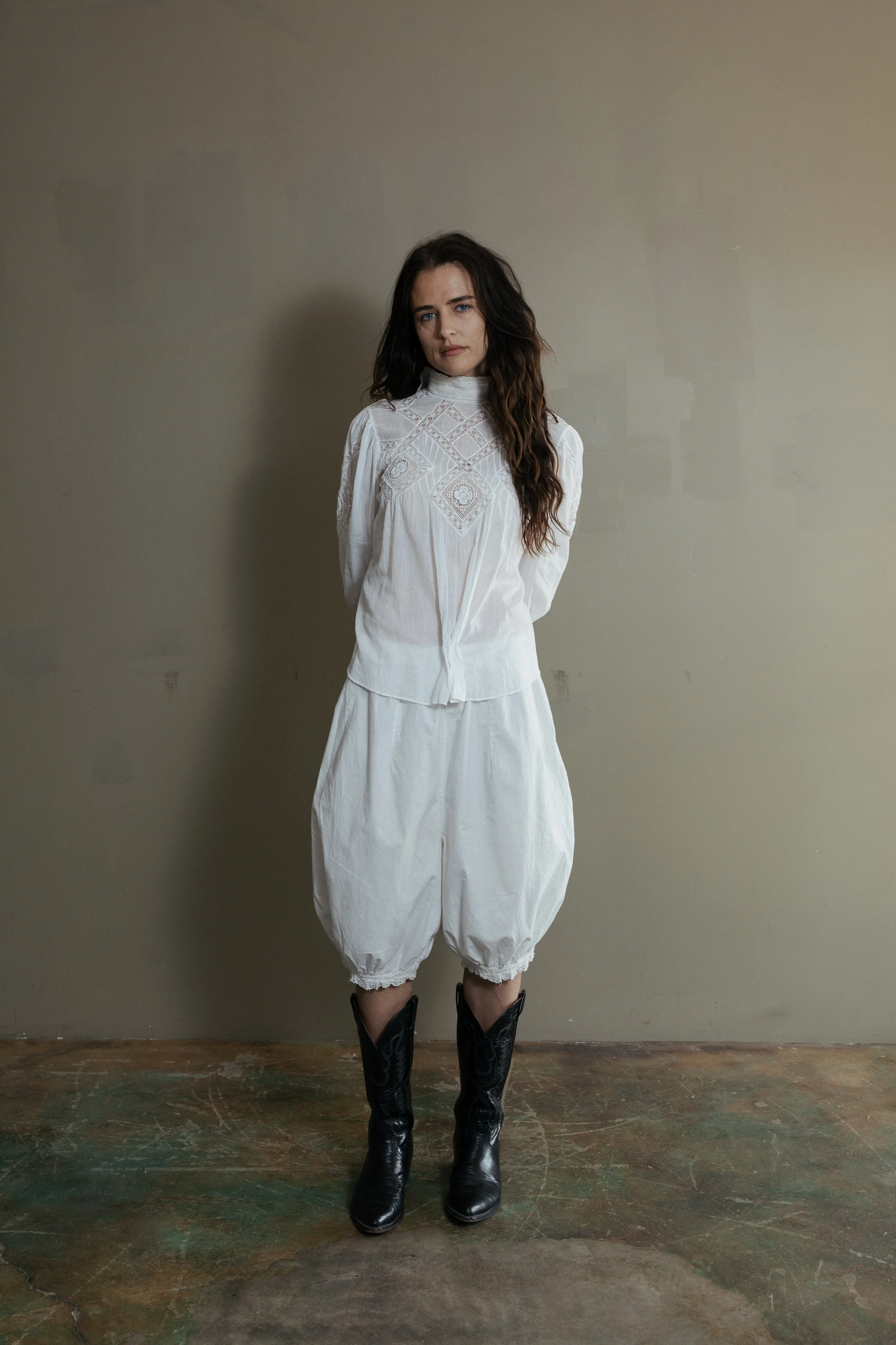 A woman standing against a plain wall, wearing a white blouse and cropped white pants with cowgirl boots.