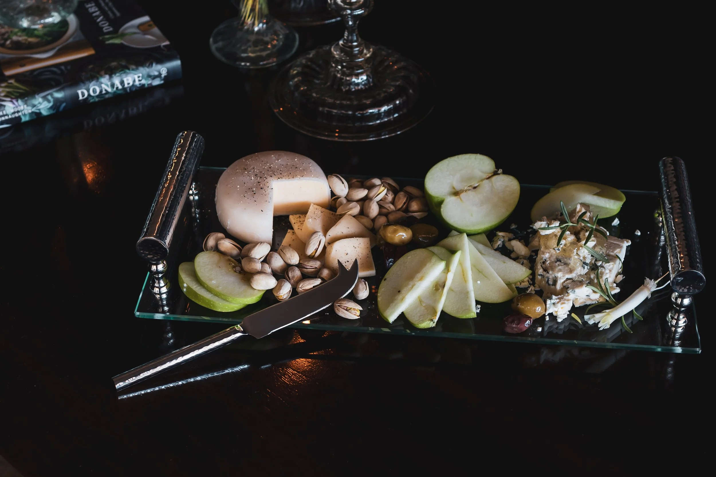 Charcuterie board with cheese, sliced green apple, pistach nuts, grapes, and a cheese knife on a black table.