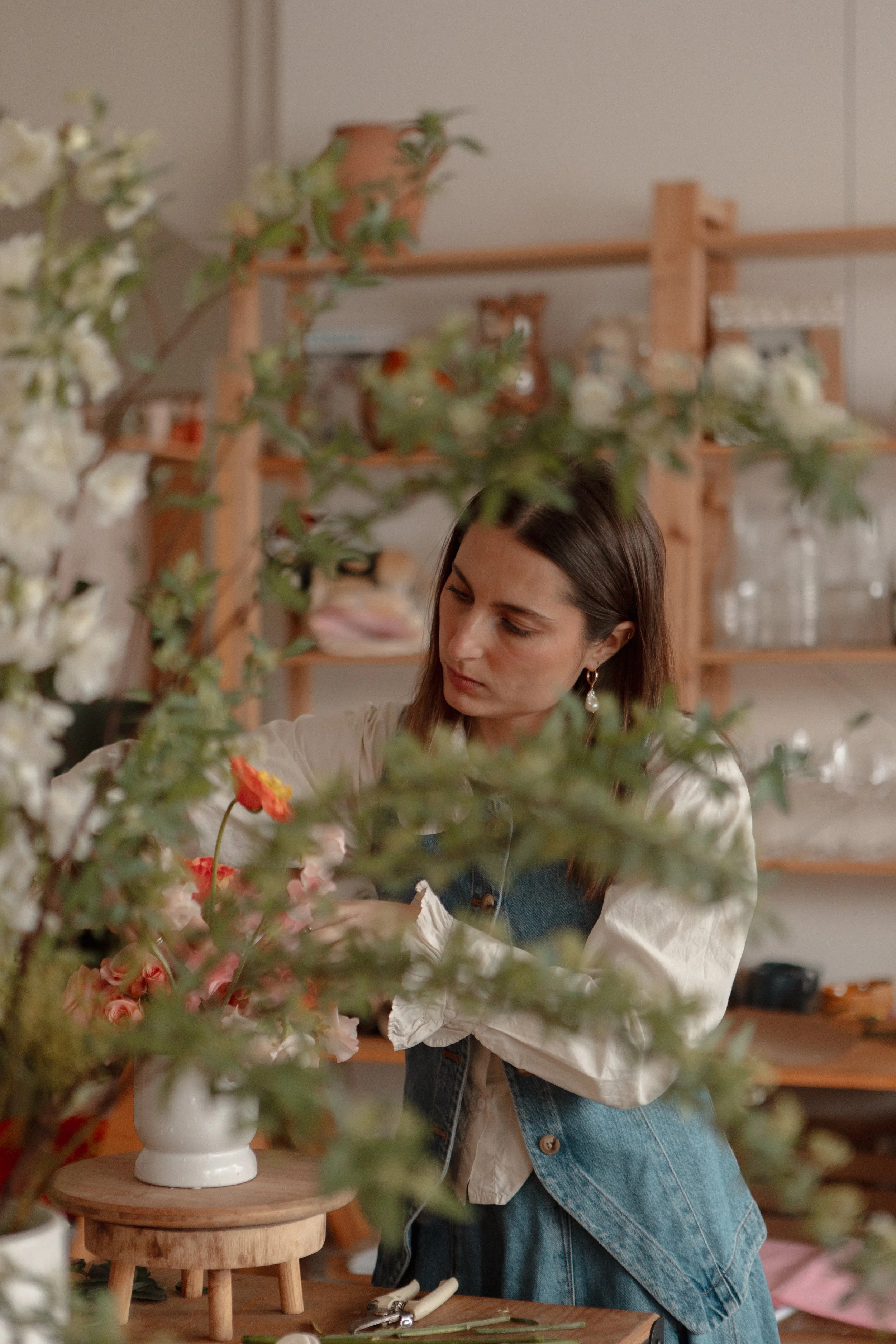 A woman arranging flowers in a white vase on a wooden table in a cozy, well-organized floral shop.