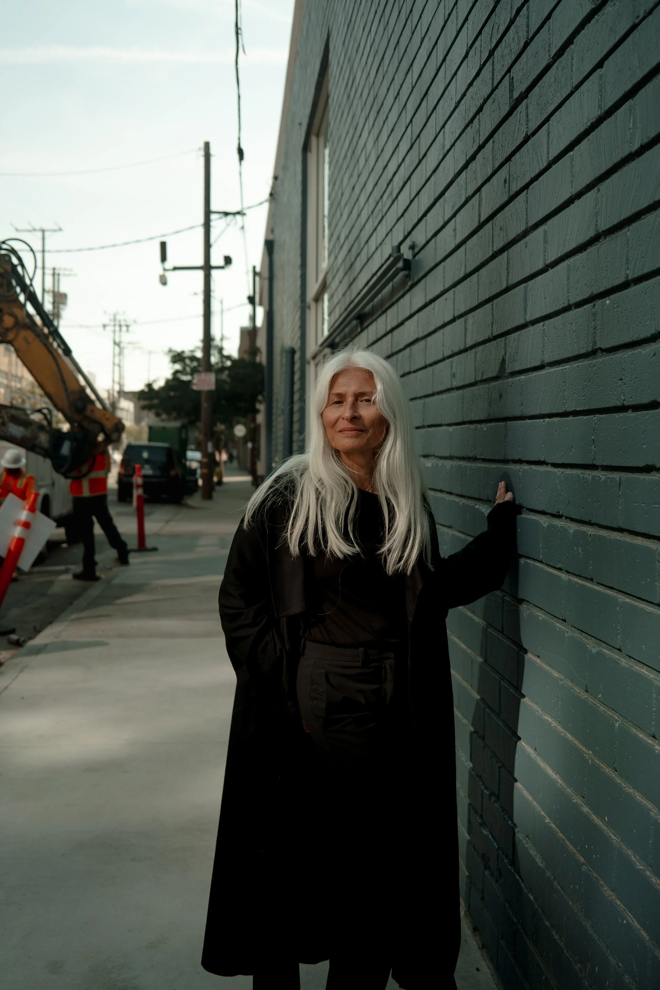 An older woman with long white hair standing against a dark green brick wall on a city sidewalk during daytime, with construction workers and equipment visible in the background.