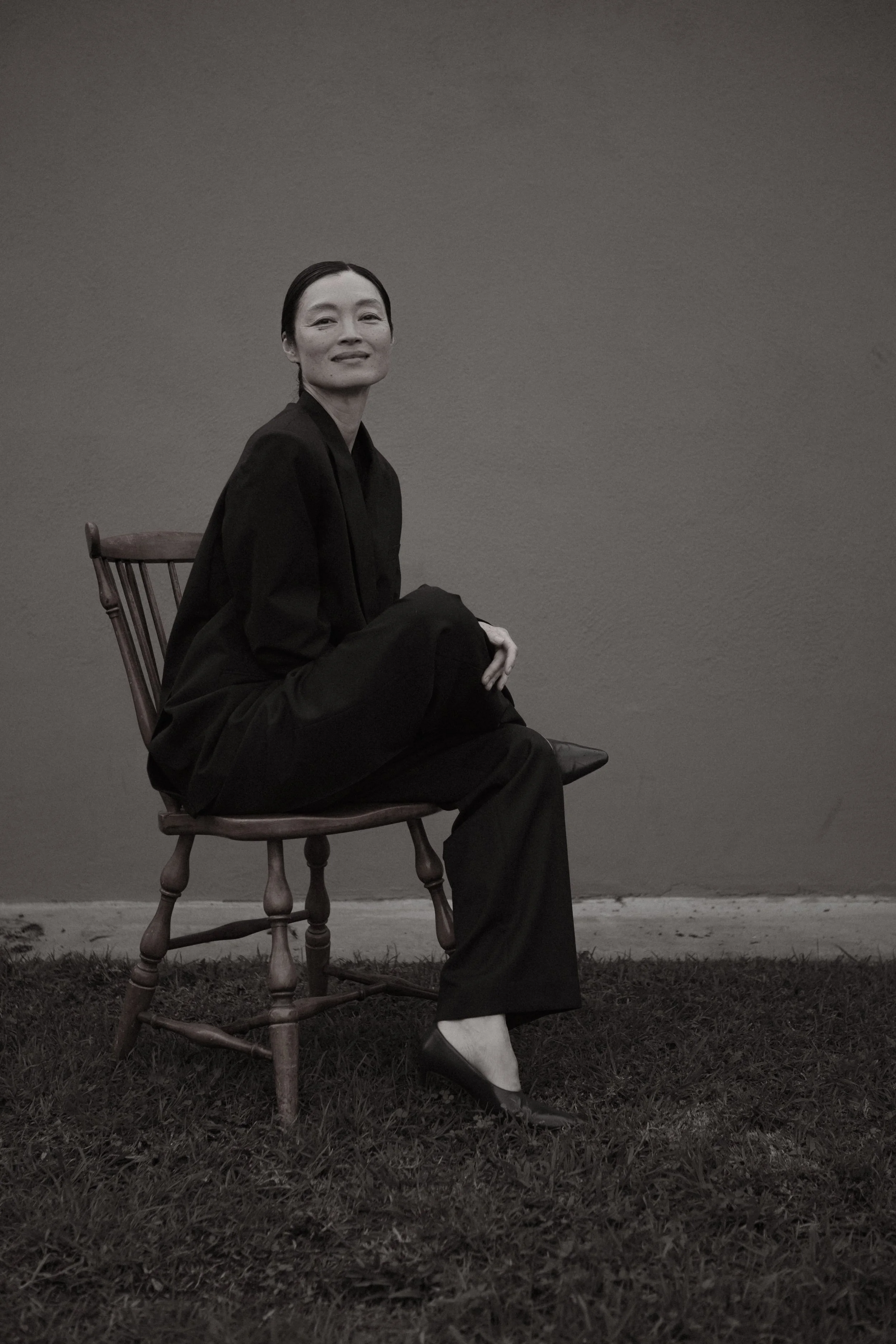 A woman in a black outfit sitting on a wooden chair against a plain wall, with grass underneath, looking at the camera.