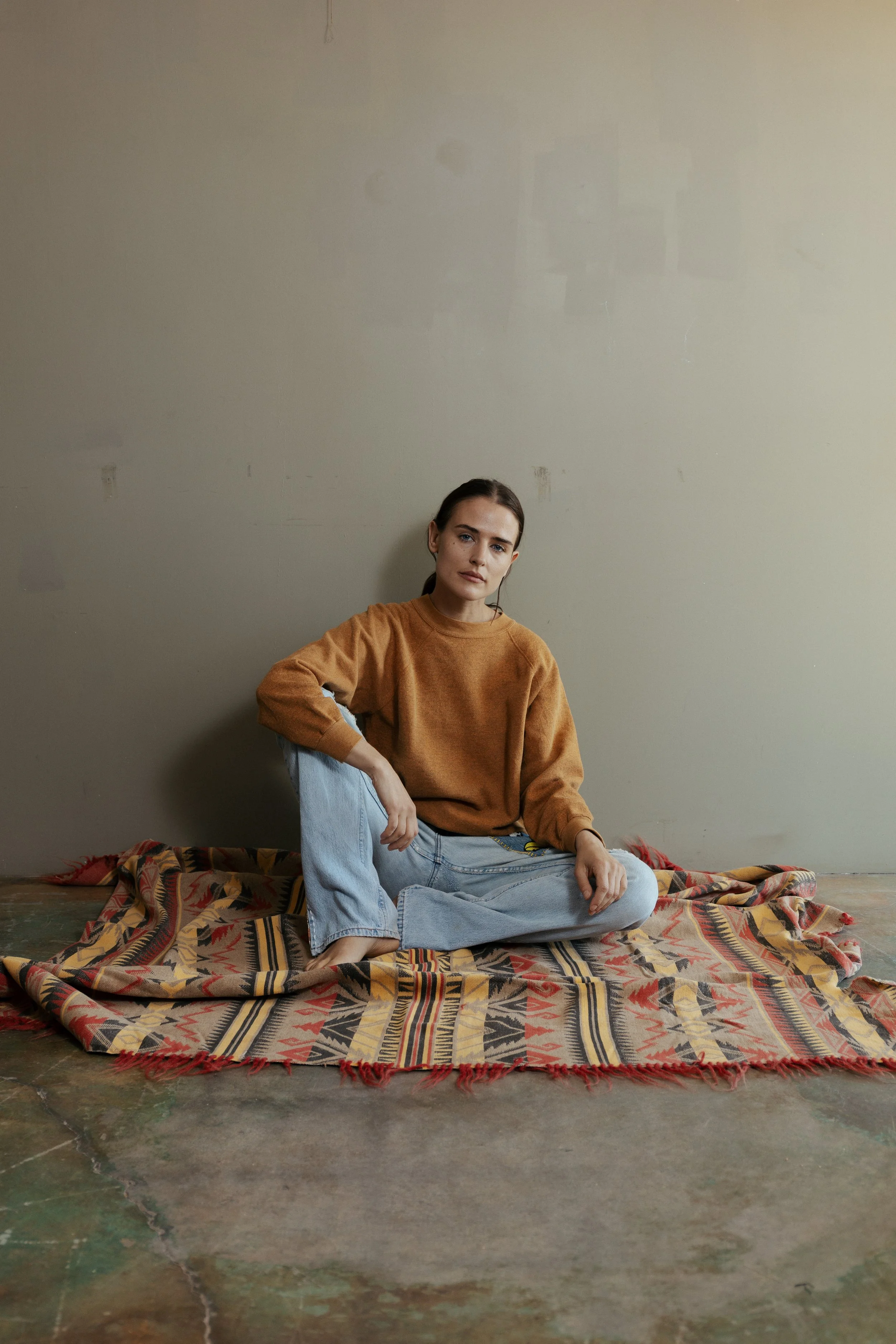 A woman sitting cross-legged on a patterned rug against a plain wall, wearing a brown sweatshirt and light blue jeans.