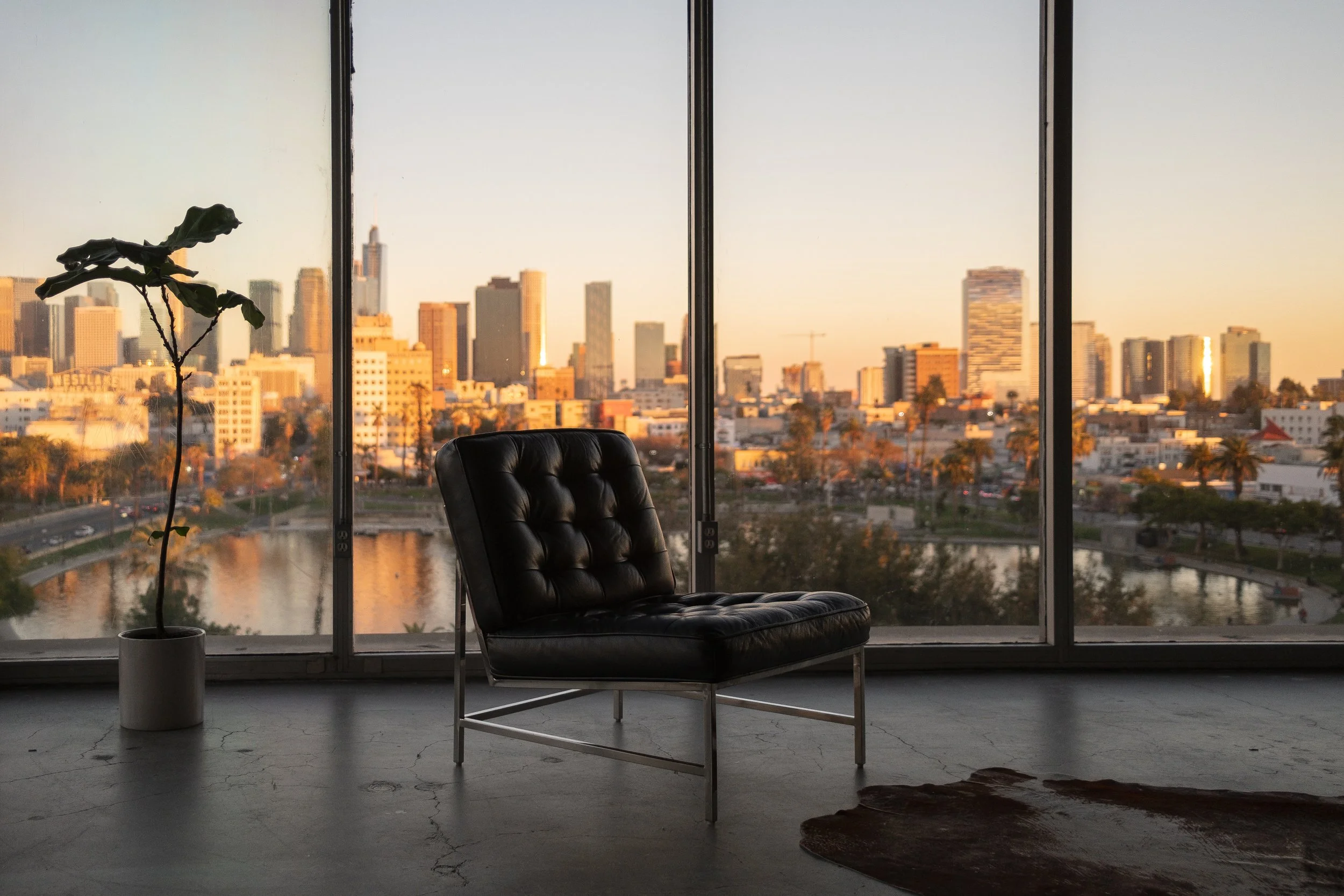 Interior of a modern apartment with a black tufted chair, a potted plant, and a large window overlooking a city skyline at sunset, with buildings, trees, and a river below.
