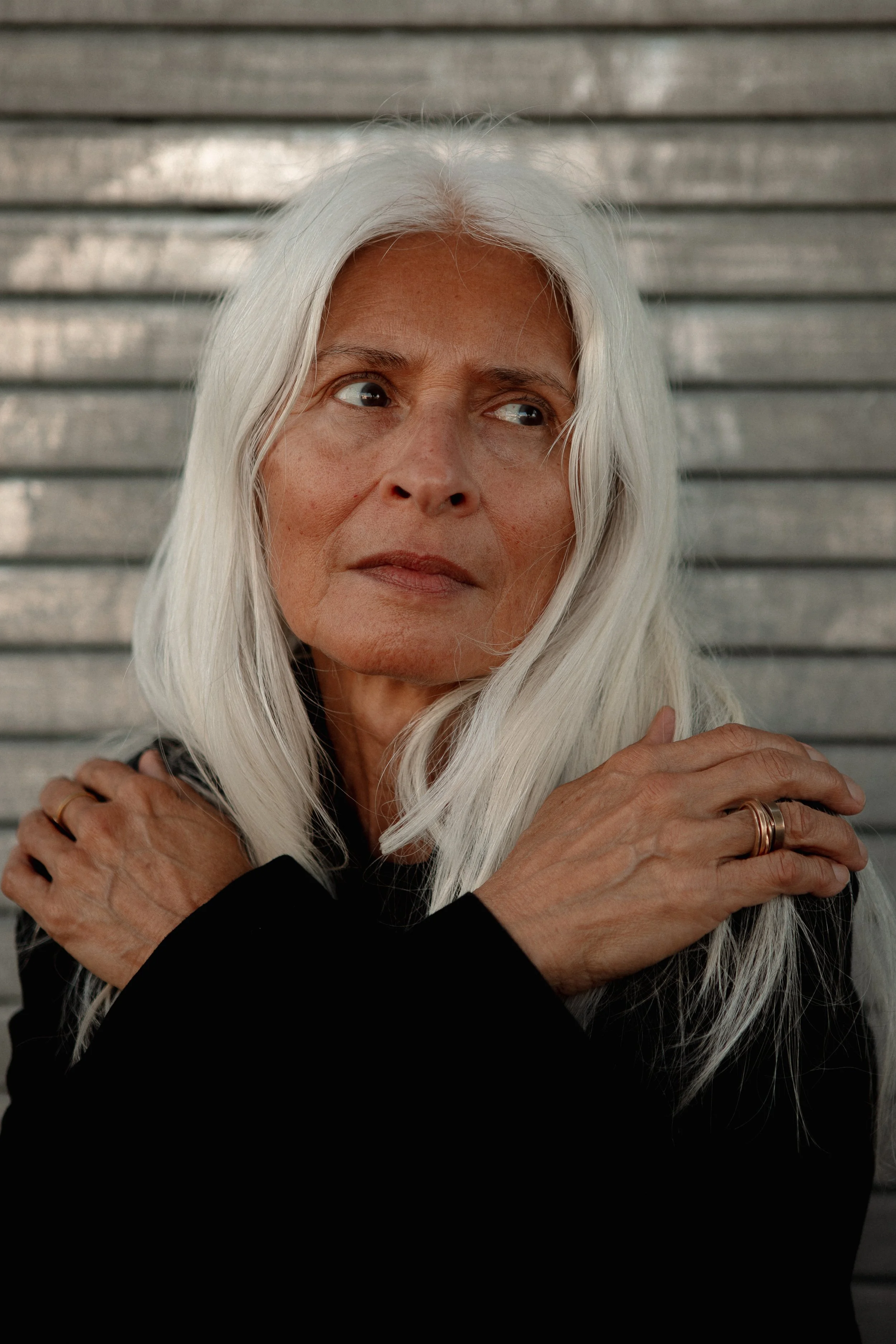 A woman with long white hair and a serious expression, looking to her right, with crossed arms and a black top, standing against a metallic background.
